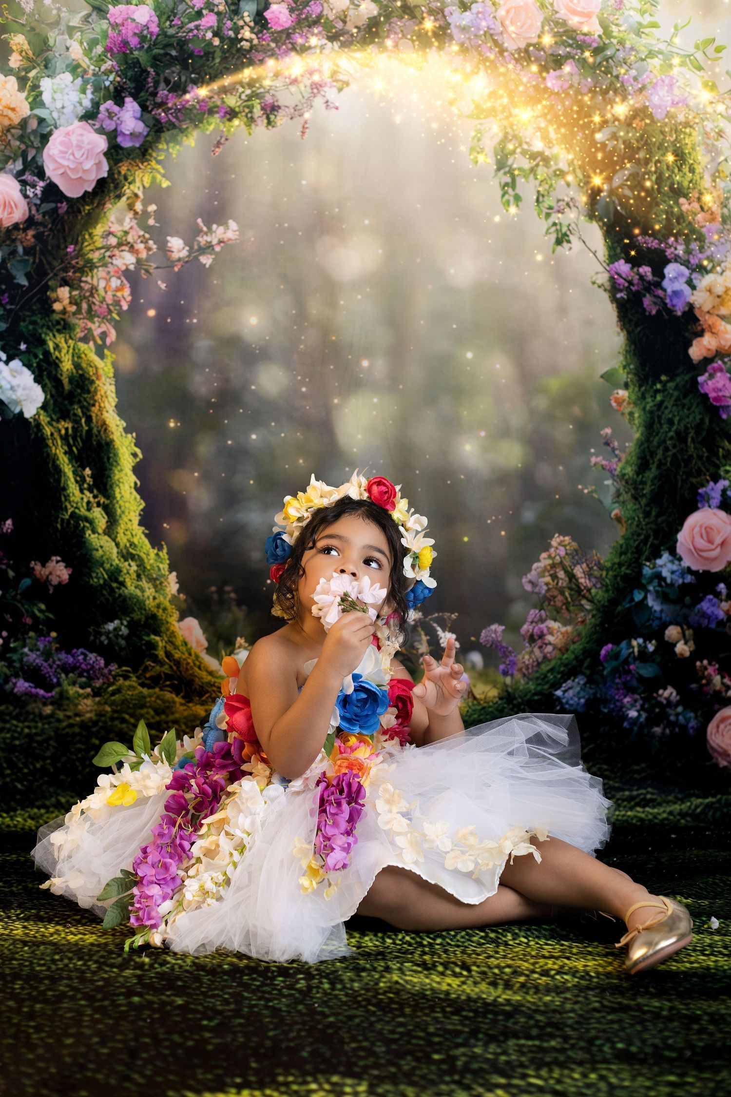 A little girl in a white tutu and floral crown sits in a magical garden arch surrounded by colorful flowers and sparkling lights.