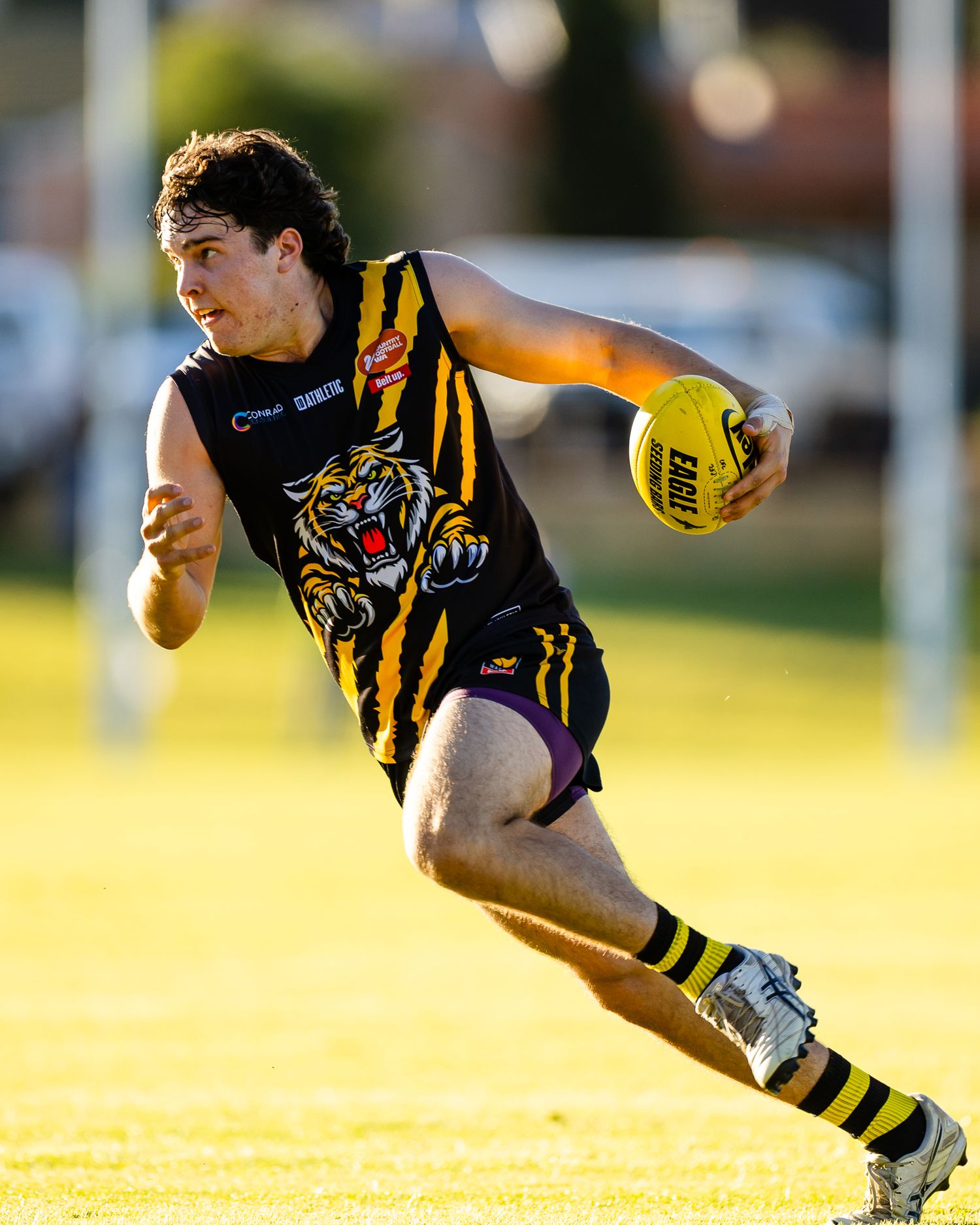 Young AFL player in tiger-striped black and gold uniform running with football during a match on green grass field.