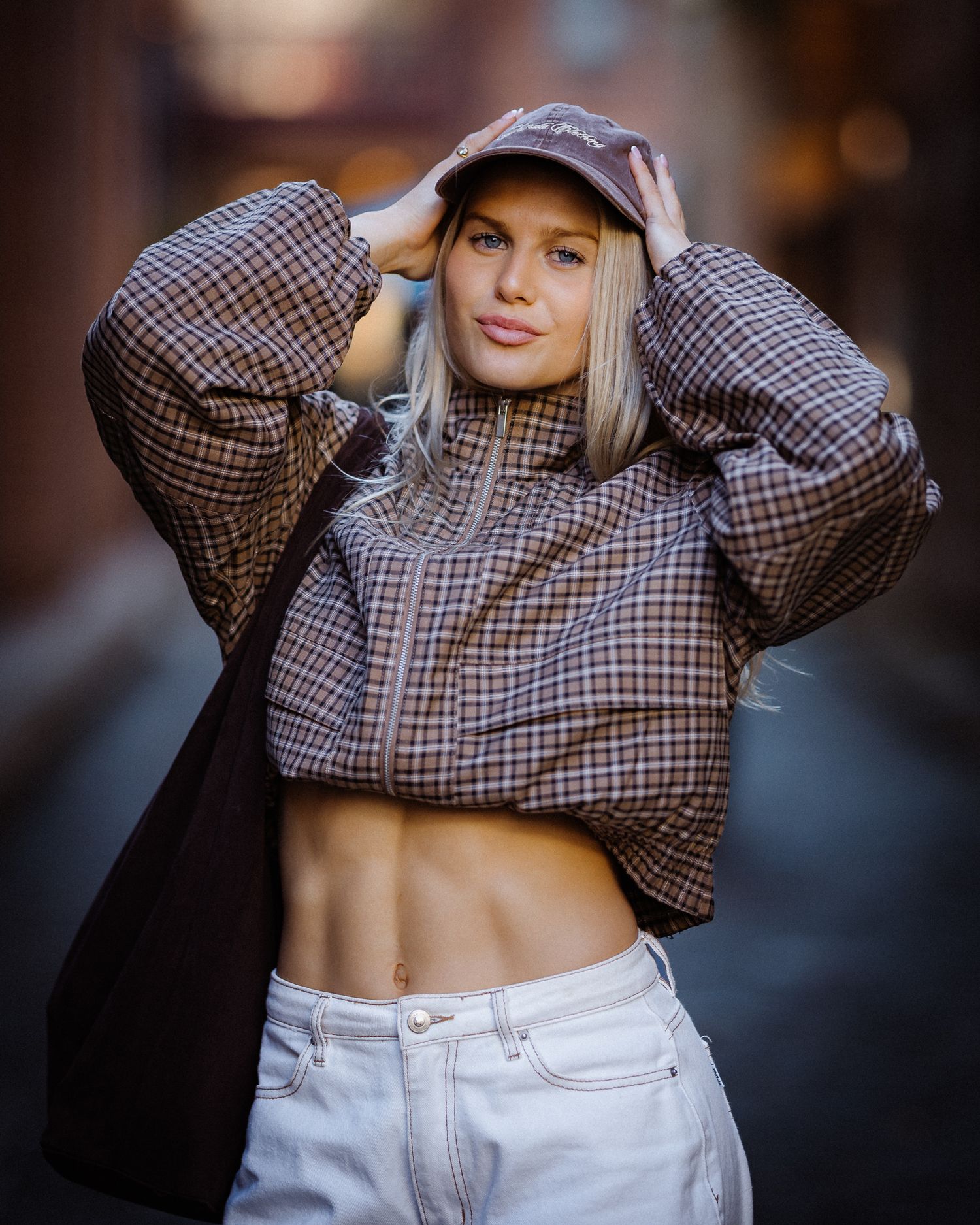 Blonde woman in plaid crop top and white jeans adjusting baseball cap in urban alley setting.