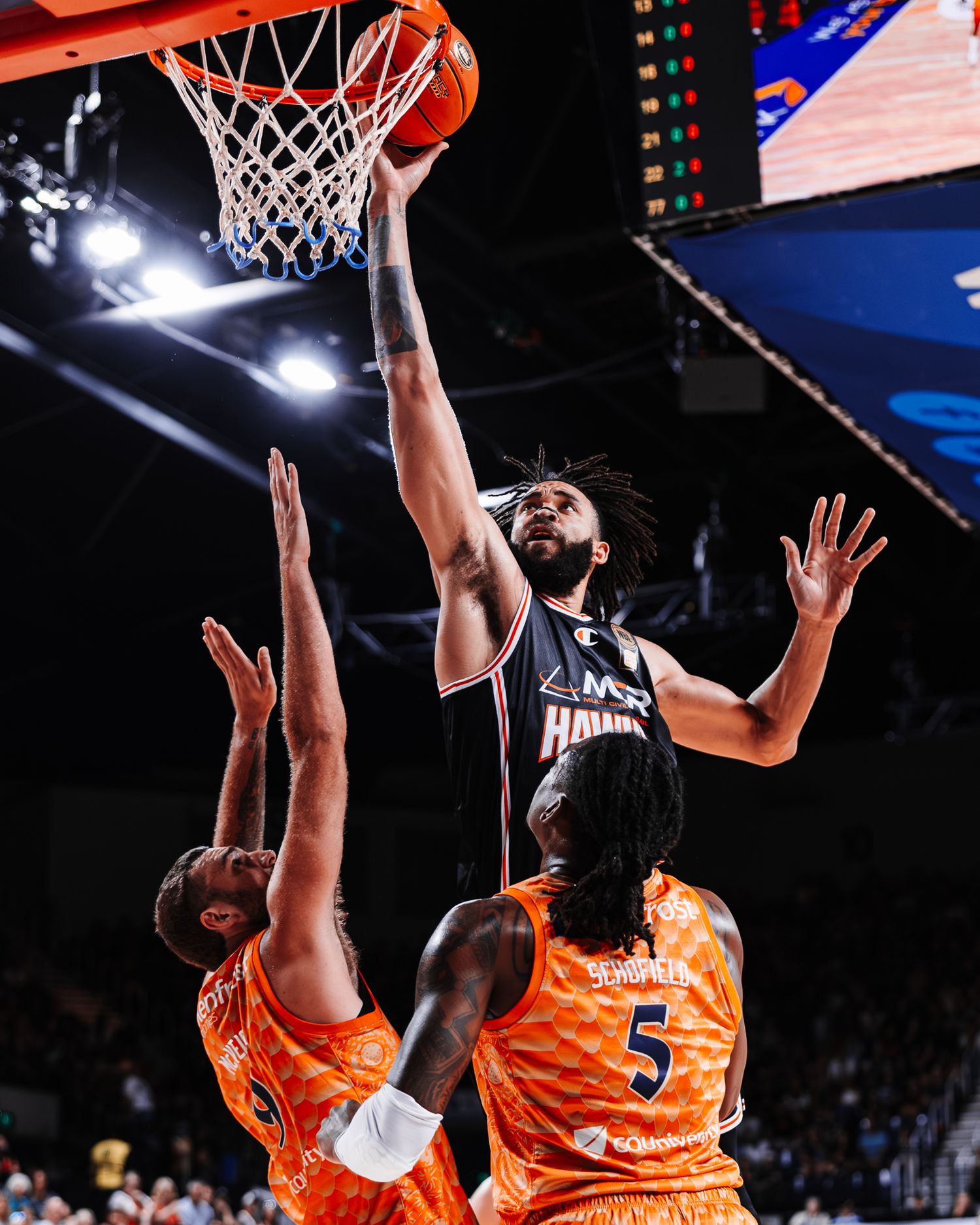 Basketball players leap for a rebound under the hoop during an intense game with orange jerseys visible.