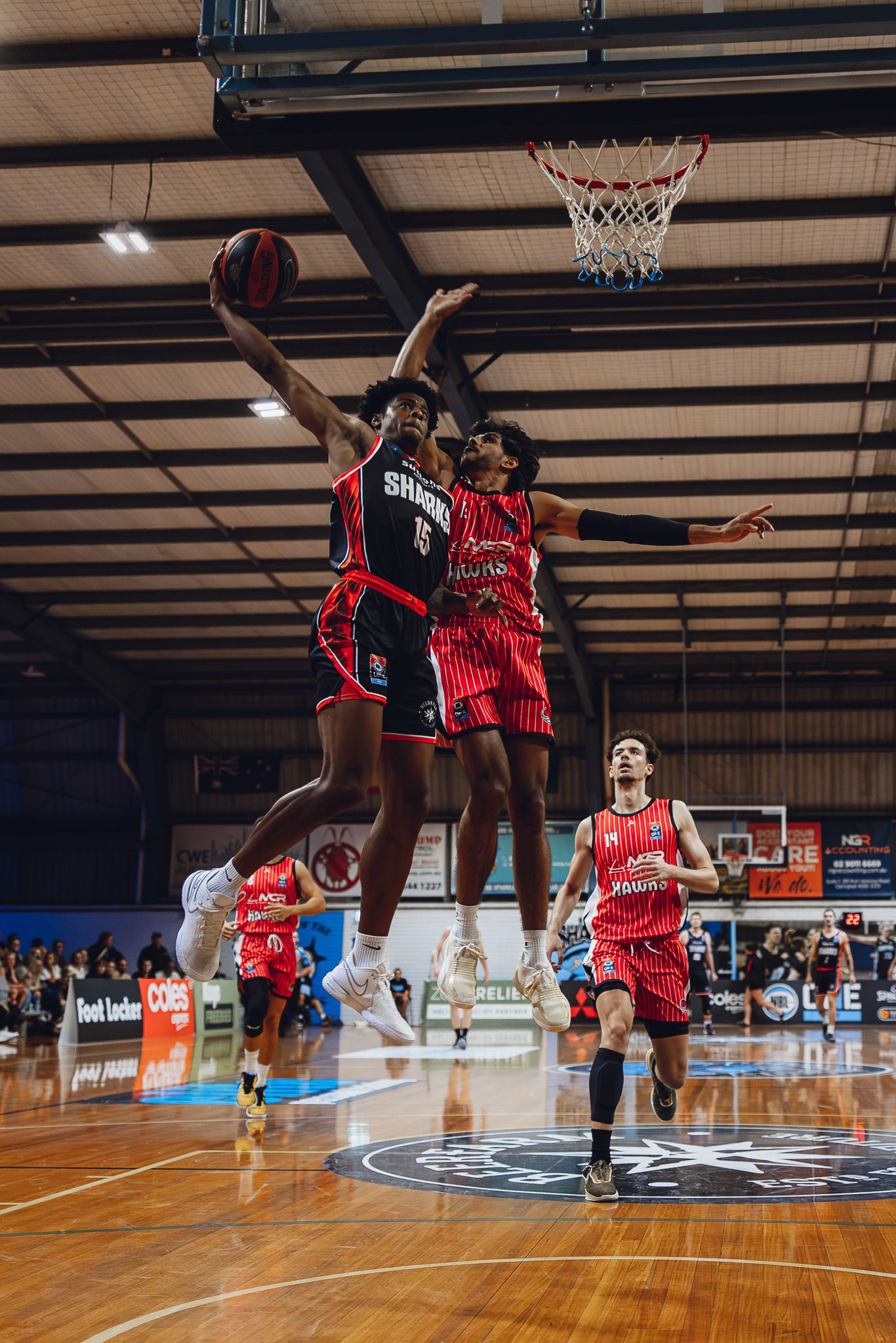 Basketball players leap high for the ball during an intense indoor game on a wooden court with spectators watching.