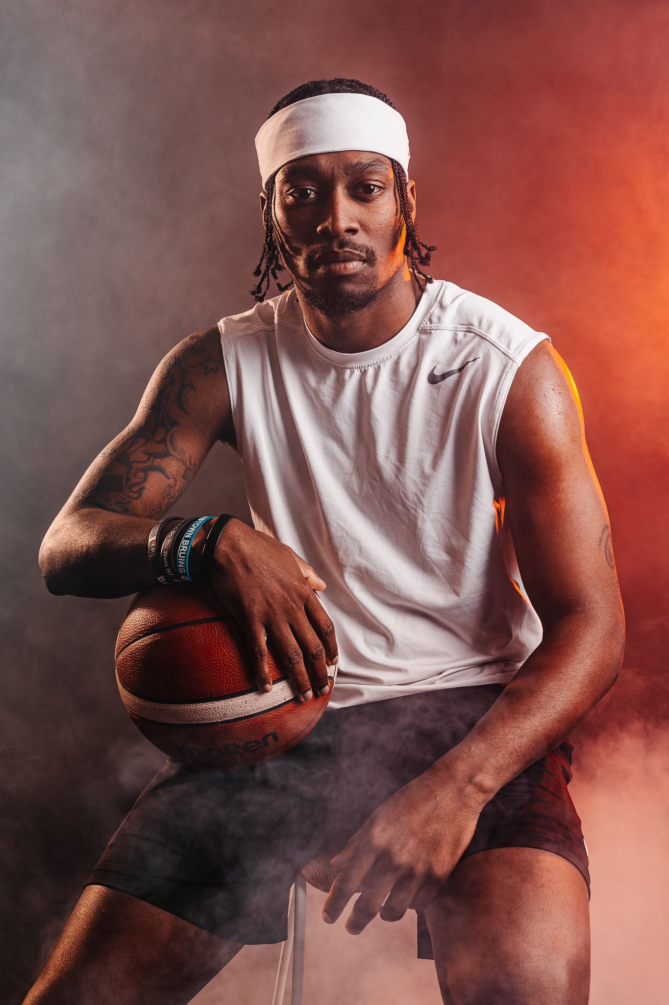 Basketball player in white sleeveless shirt poses with ball in dramatic red-lit studio setting.
