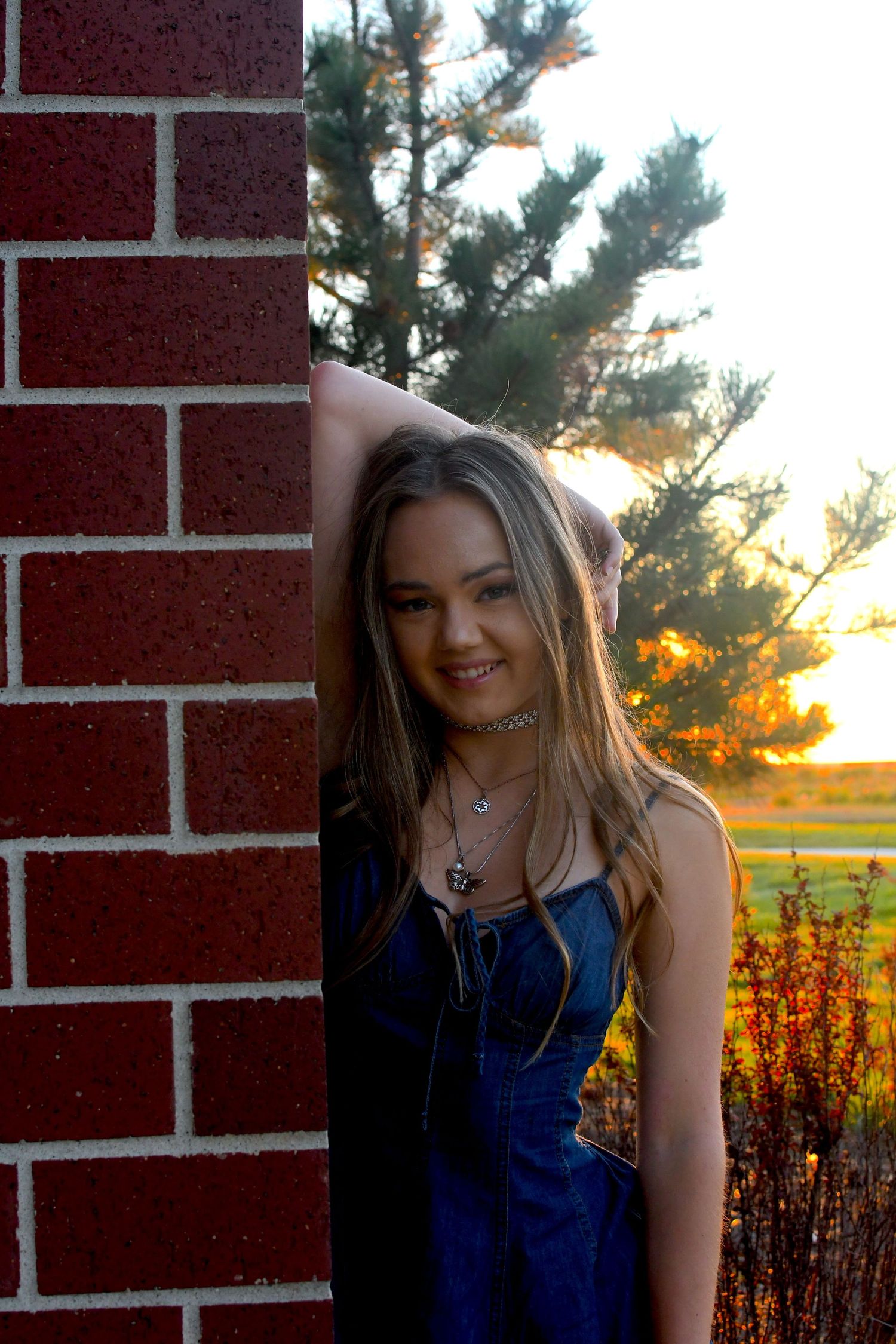 Person in blue dress poses against brick wall during golden sunset with flowers and trees in background.