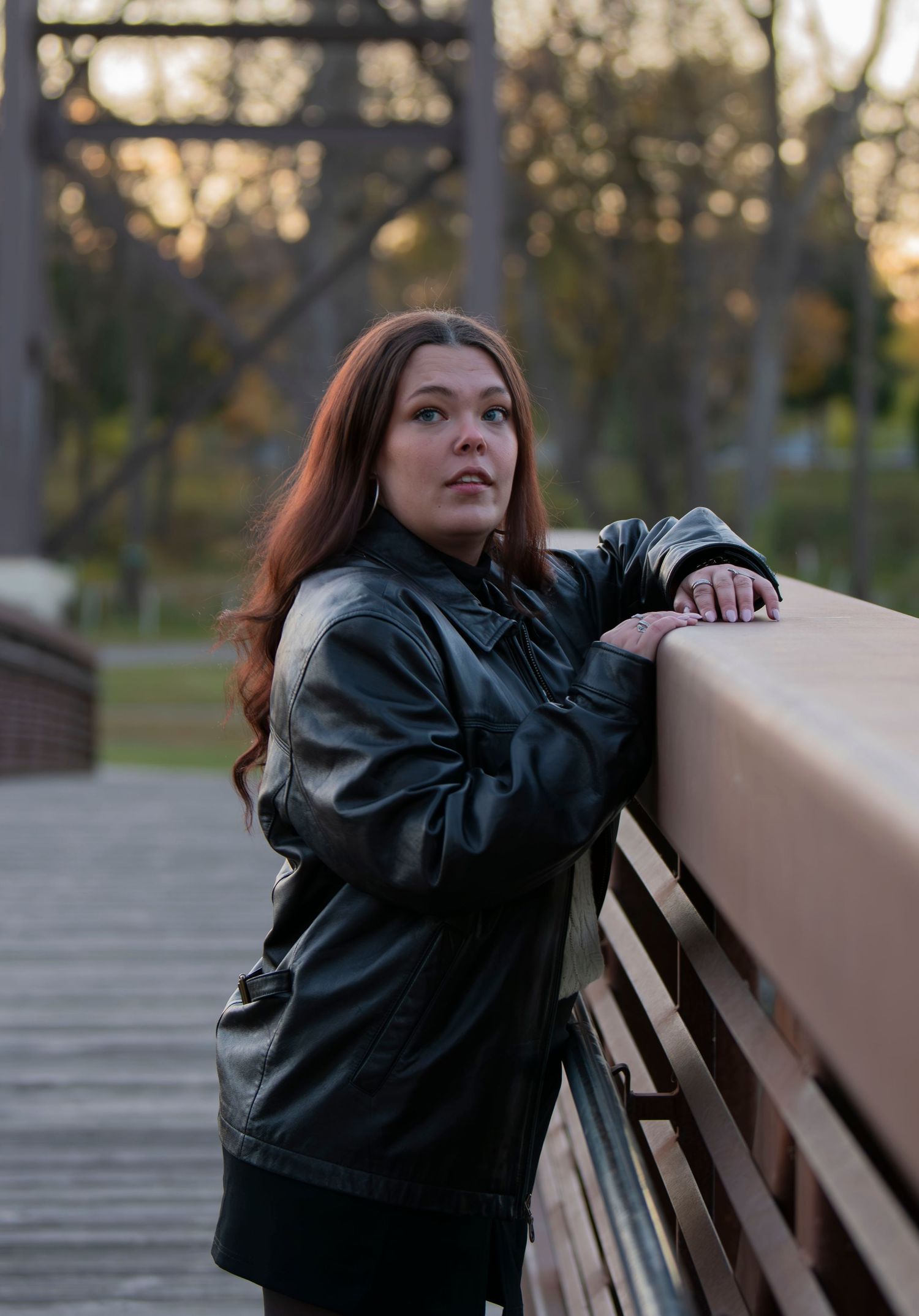 A person in a black leather jacket leans against a railing on a bridge at sunset.