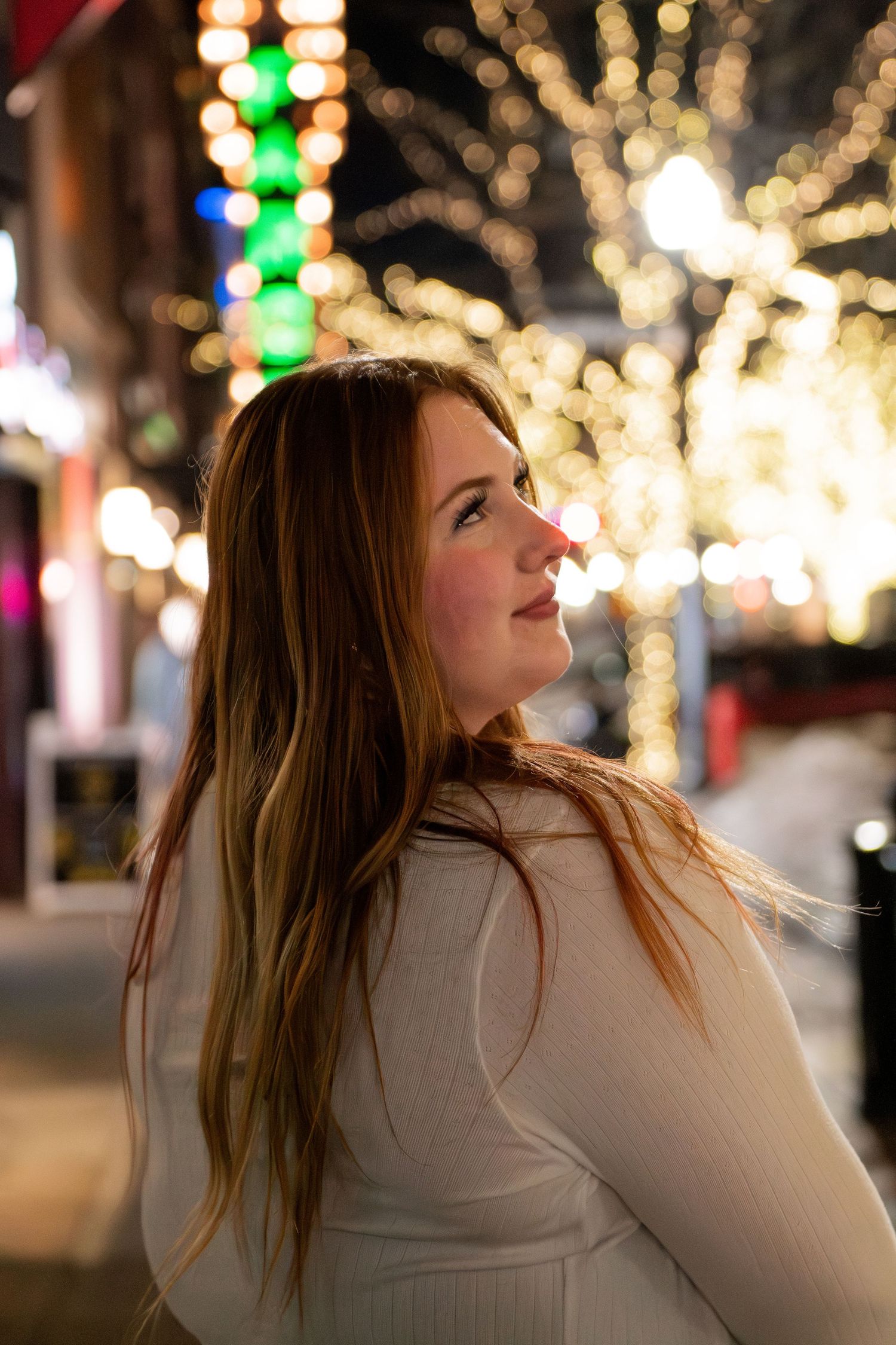 Woman in white sweater looks up at holiday lights glowing in bokeh against dark night sky.