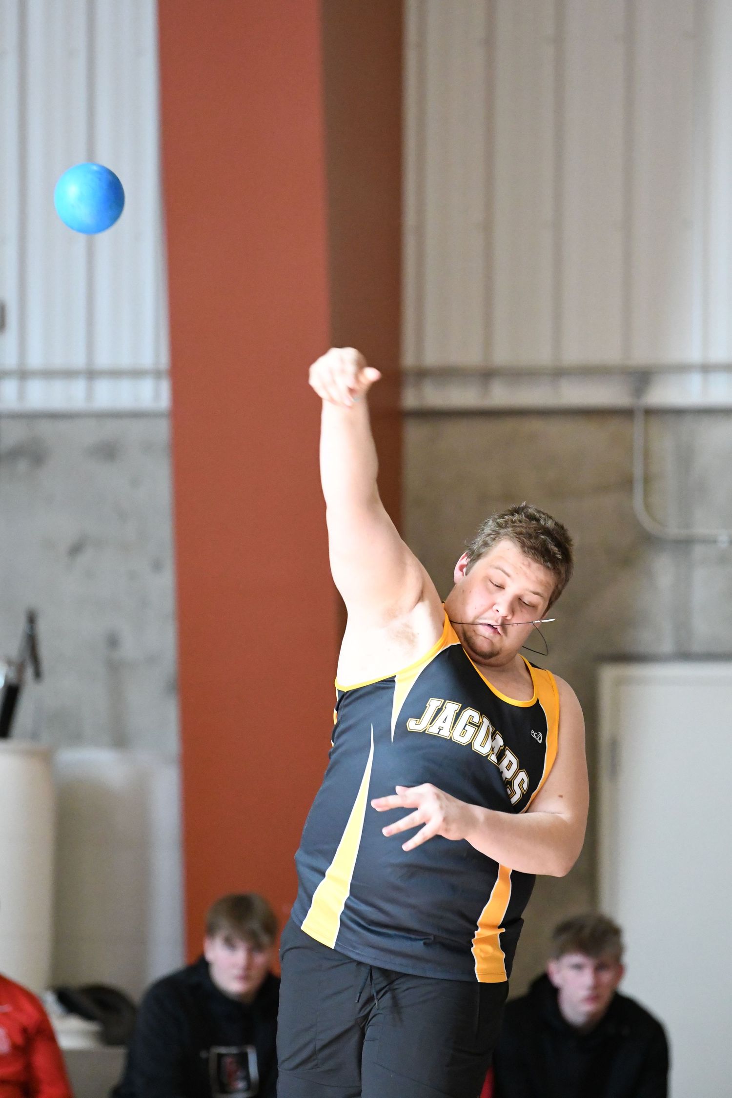 An athlete in black and gold uniform throws a shot put during an indoor track competition.