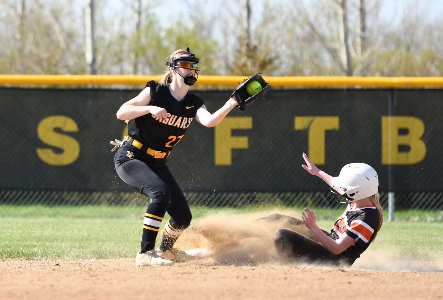 Player slides into base as softball game action unfolds on dirt infield.