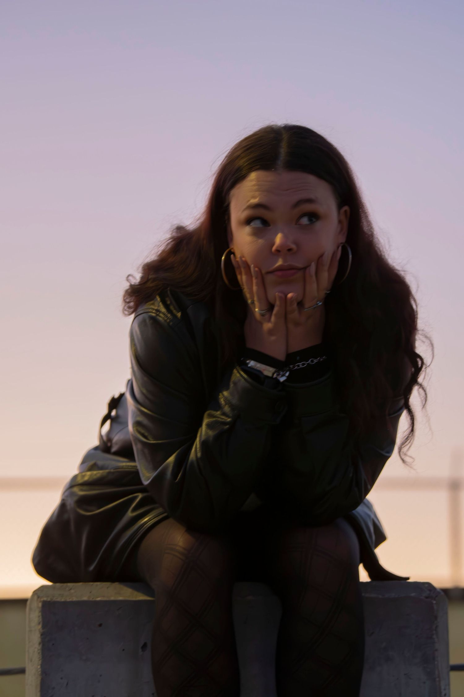 A person in dark clothing sits thoughtfully on a ledge against a dusky purple sky.