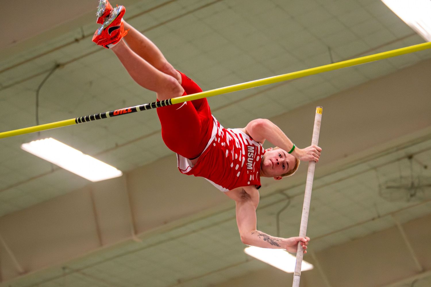 Athlete in red uniform performing pole vault technique at indoor track competition.