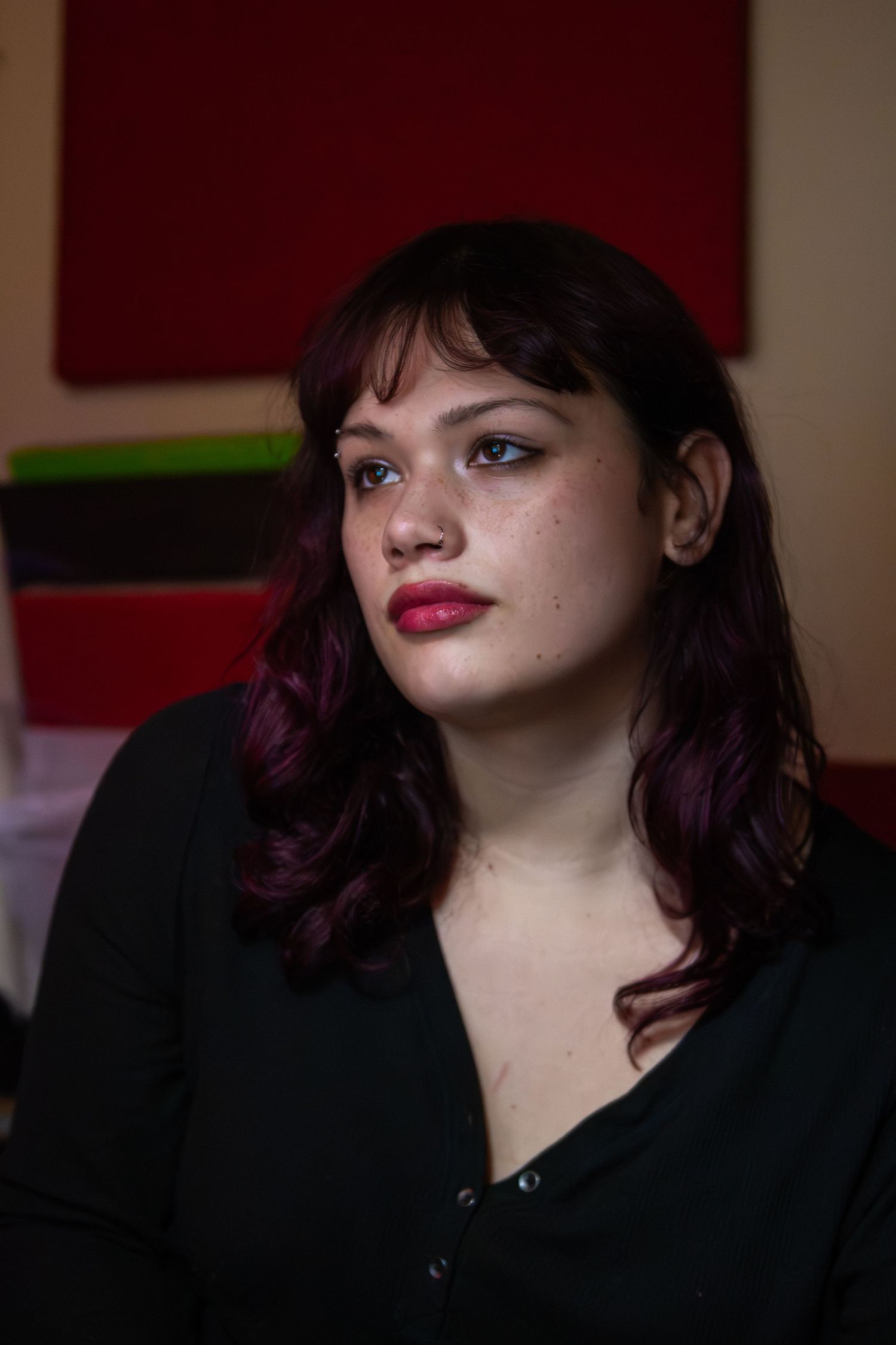 Portrait of someone with purple-tinted hair wearing red lipstick and a black top against a dark red background.