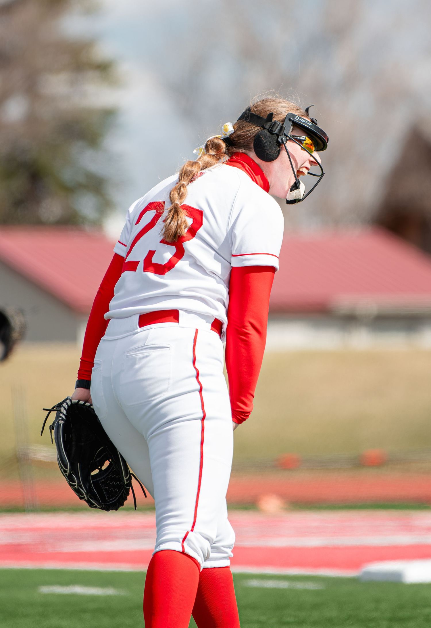 Female softball pitcher #23 in red and white uniform with batting helmet and glove on outdoor field.