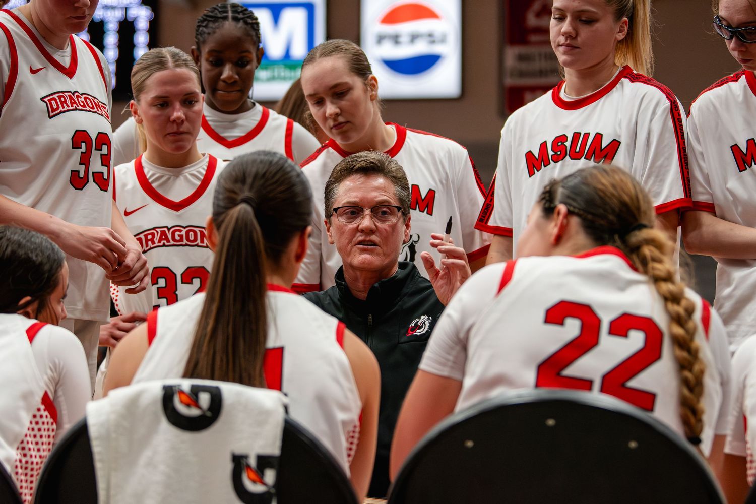 Basketball coach huddles with team wearing white and red uniforms during timeout at competitive game.