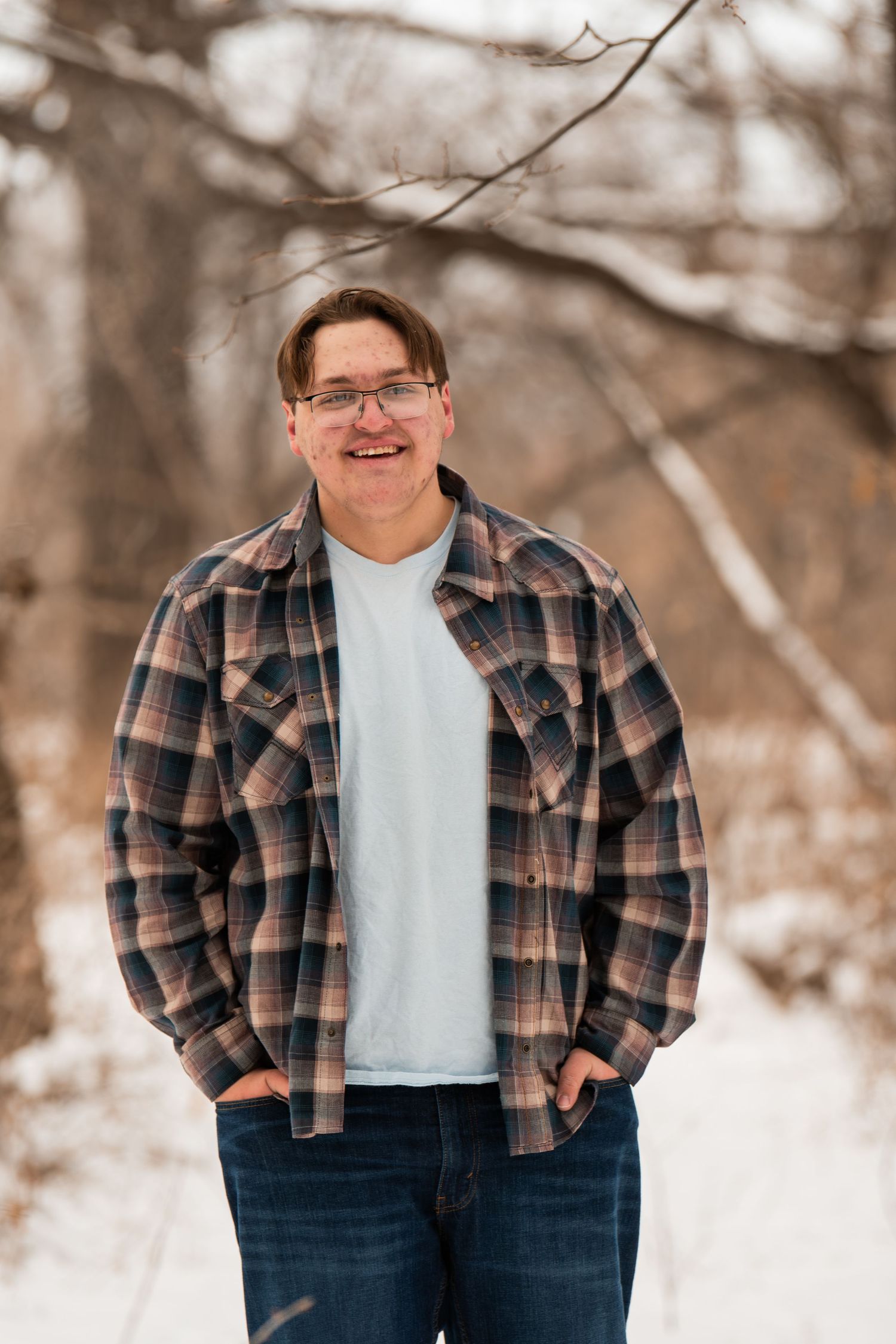 Person wearing a plaid flannel shirt and jeans poses outdoors in a winter setting with snowy trees in the background.
