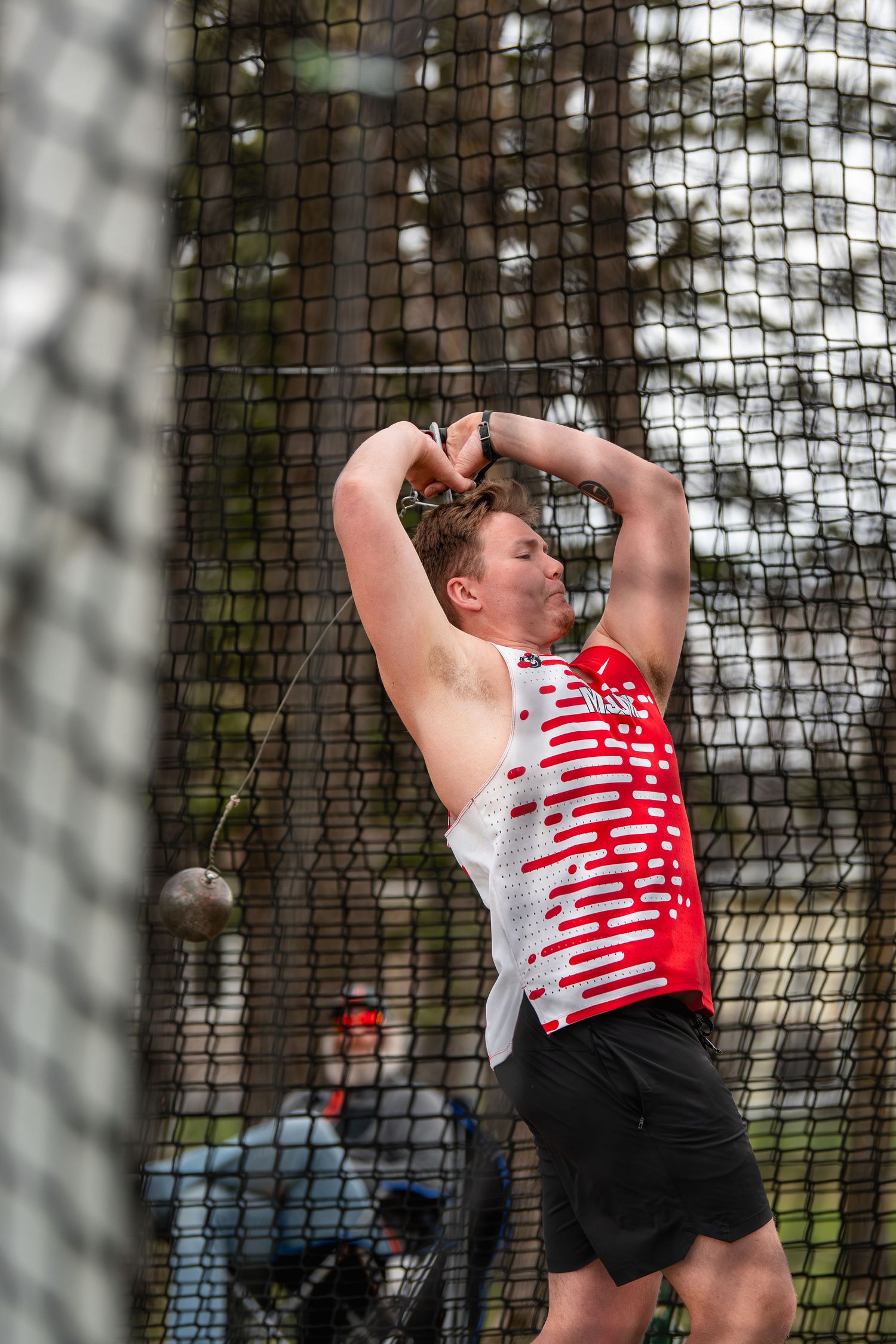 Male athlete in red uniform throws hammer at track and field competition, captured mid-spin behind safety netting.