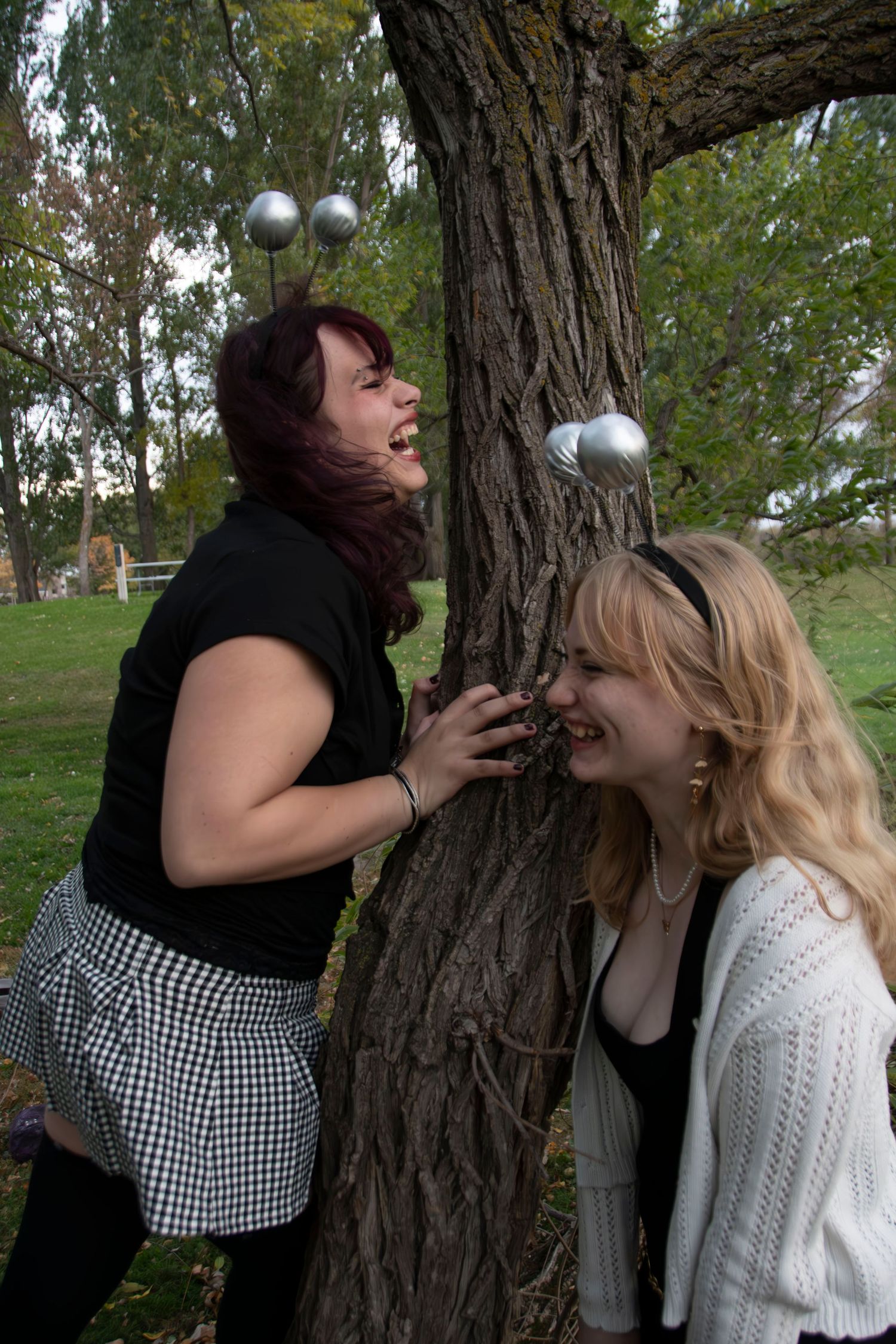 Two people playfully pose around a large tree trunk with silver ornaments in a park setting.