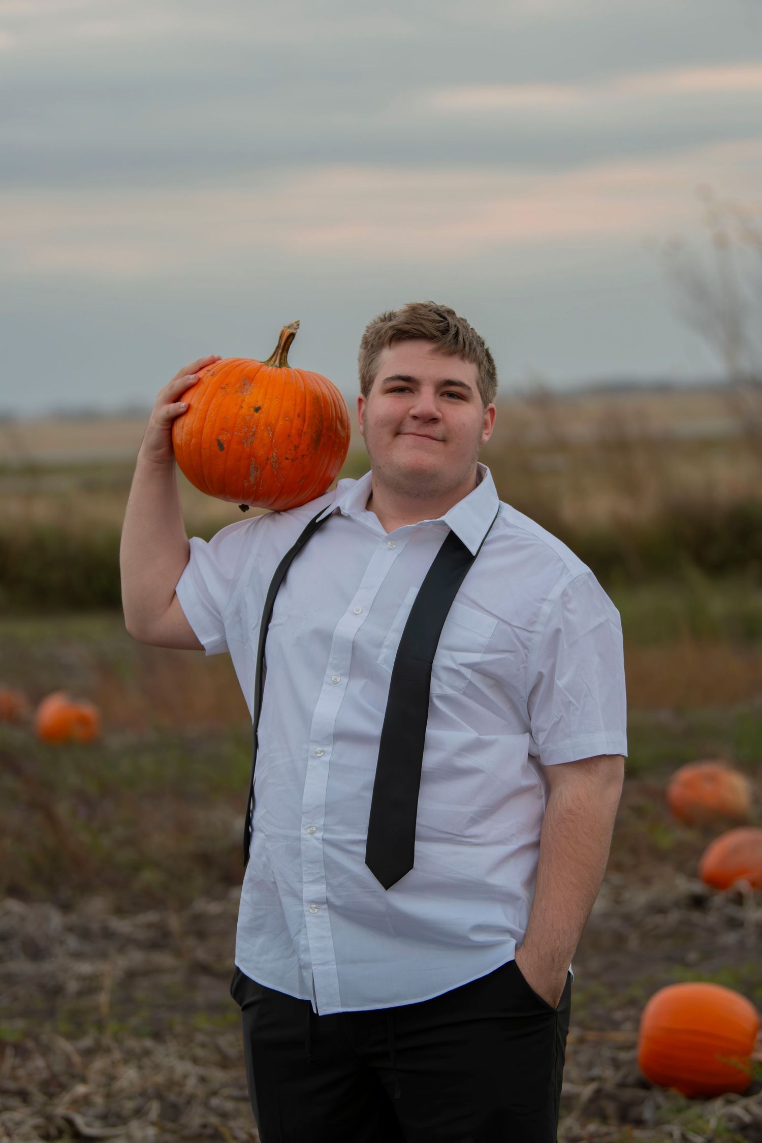 A person in a white dress shirt and black tie poses playfully in a pumpkin patch during autumn.