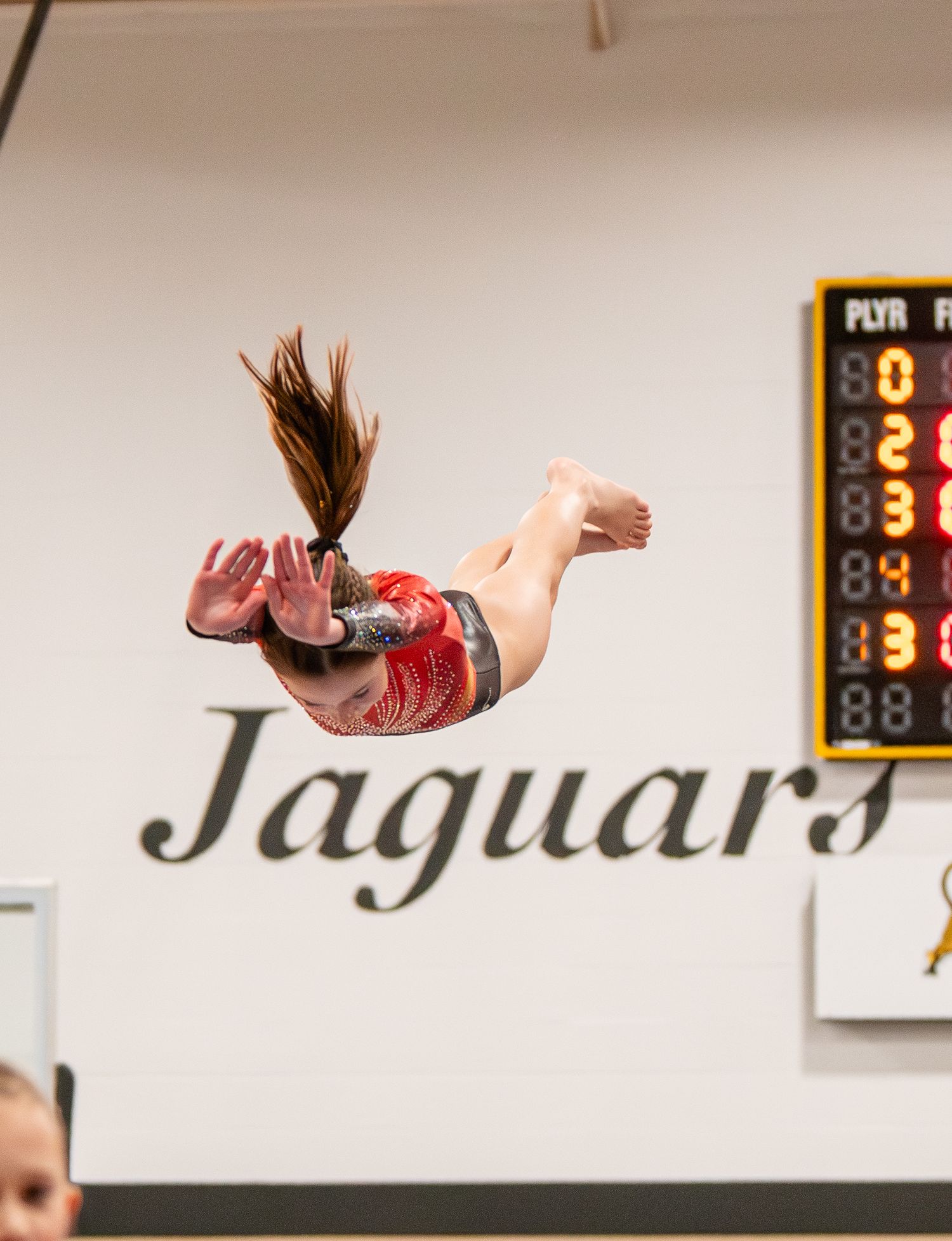 A gymnast performs a graceful split leap during a floor routine with a scoreboard visible in the background.