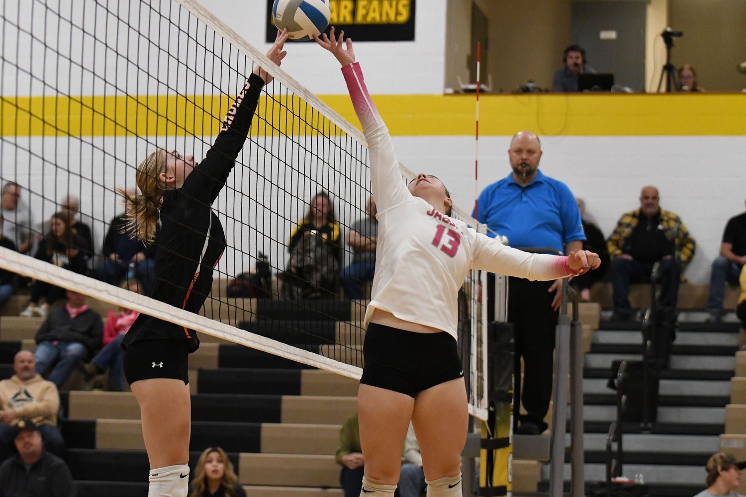 Athletes competing at the net during an indoor volleyball match with spectators watching from the stands.