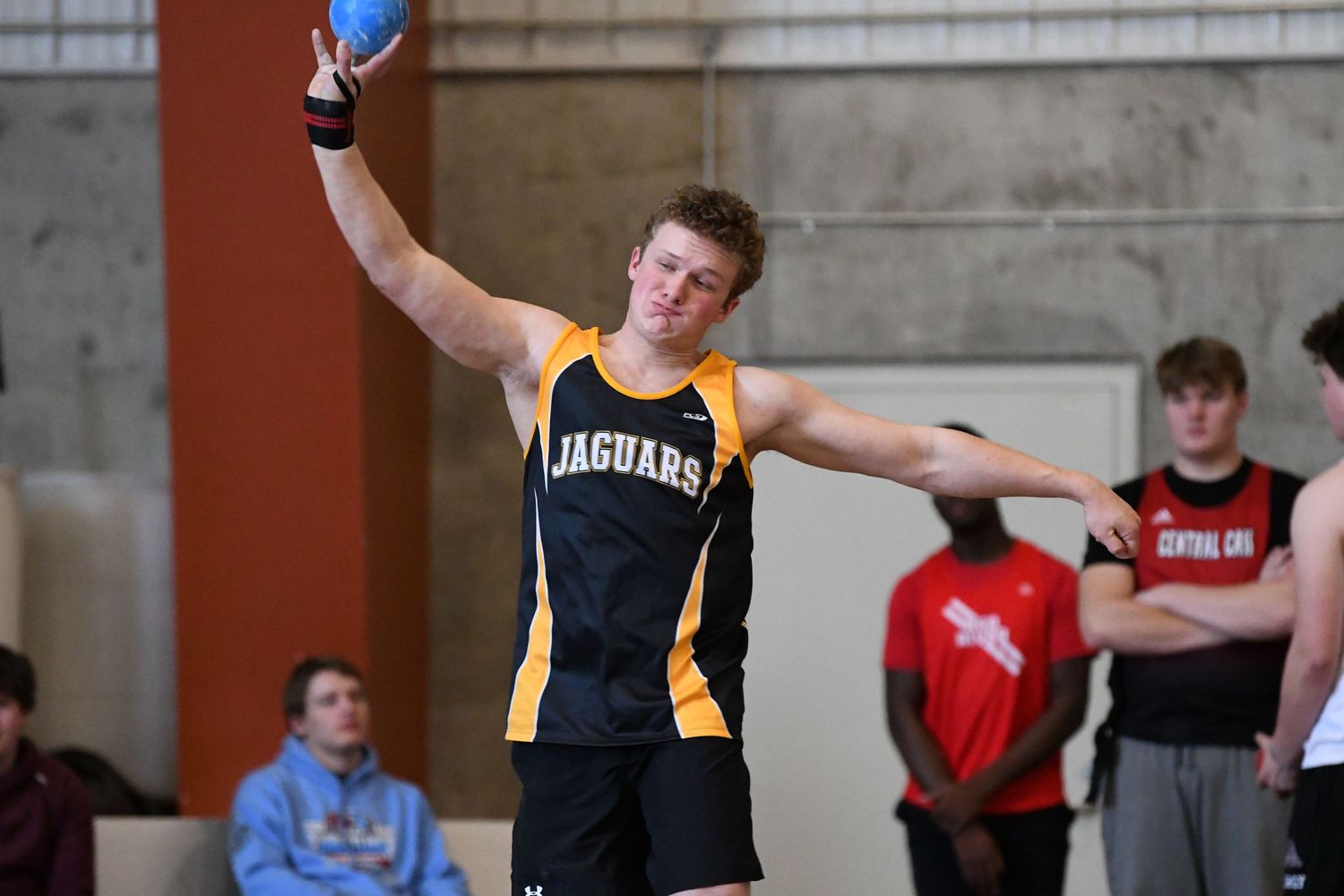 Track athlete in black and gold uniform completes shot put throw at indoor competition.