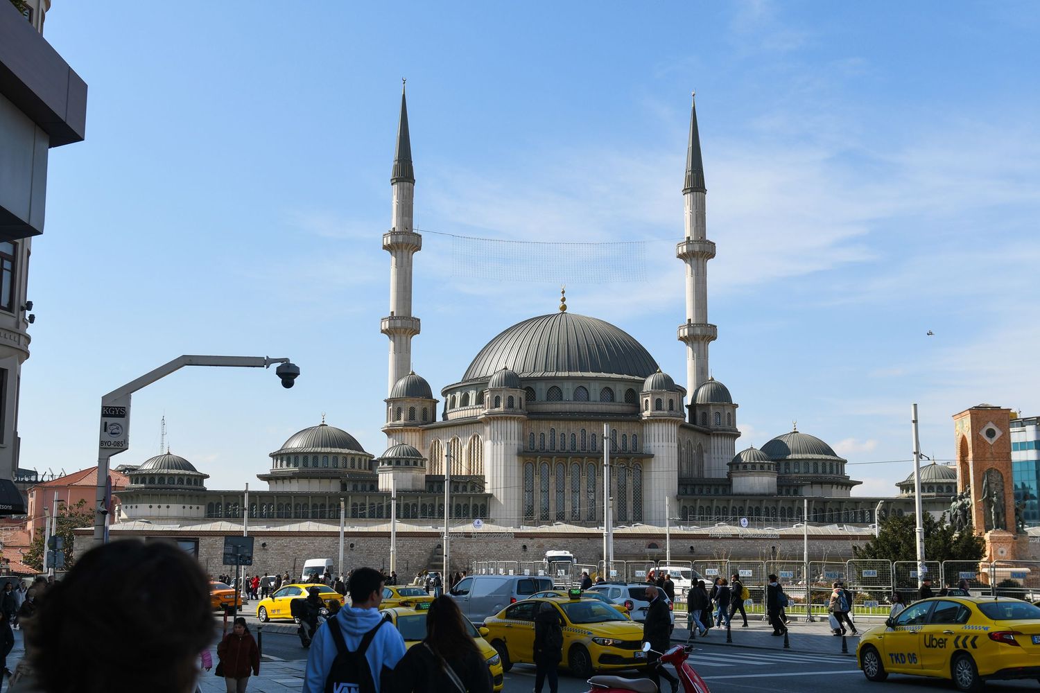 A large mosque with twin minarets and domed roof stands prominently in an urban setting with yellow taxis parked outside.