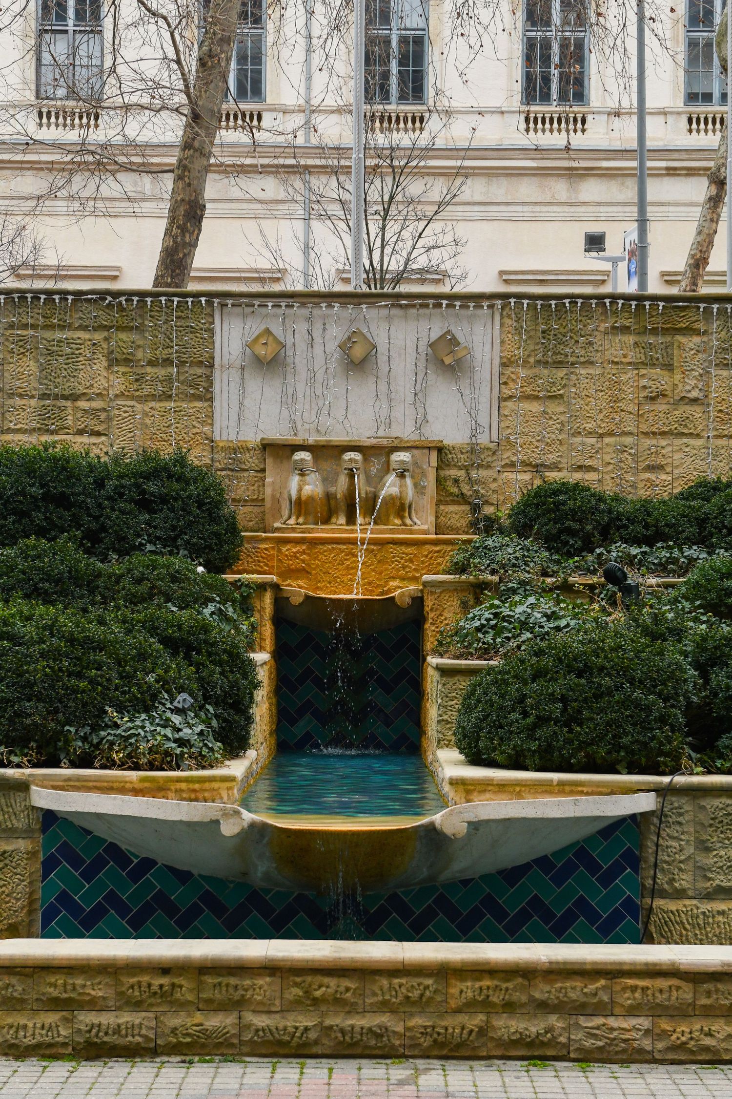 Ornate stone fountain with teal blue water and gold accents in a formal garden setting.