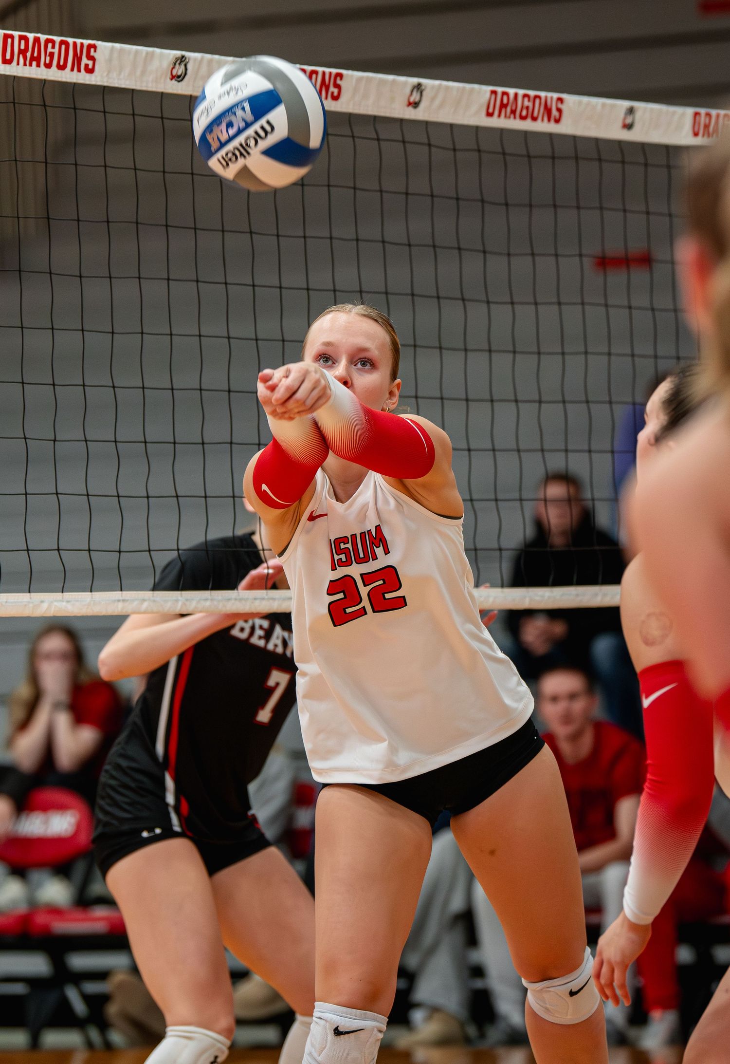 ISUM #22 volleyball player performs a forearm pass during a competitive match in front of a net with Dragons signage.