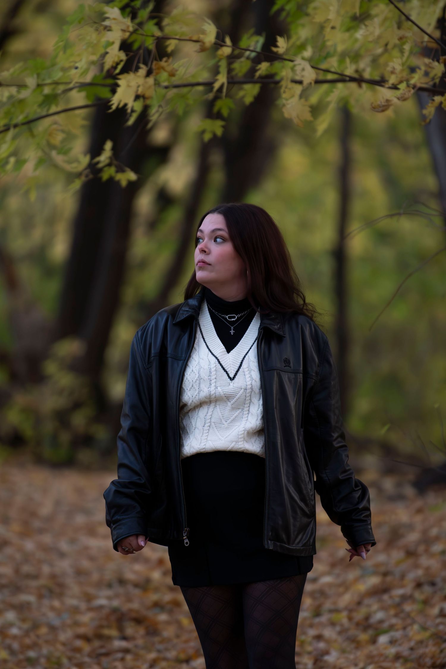 Person wearing black leather jacket and white sweater walks through autumn forest with fallen leaves.