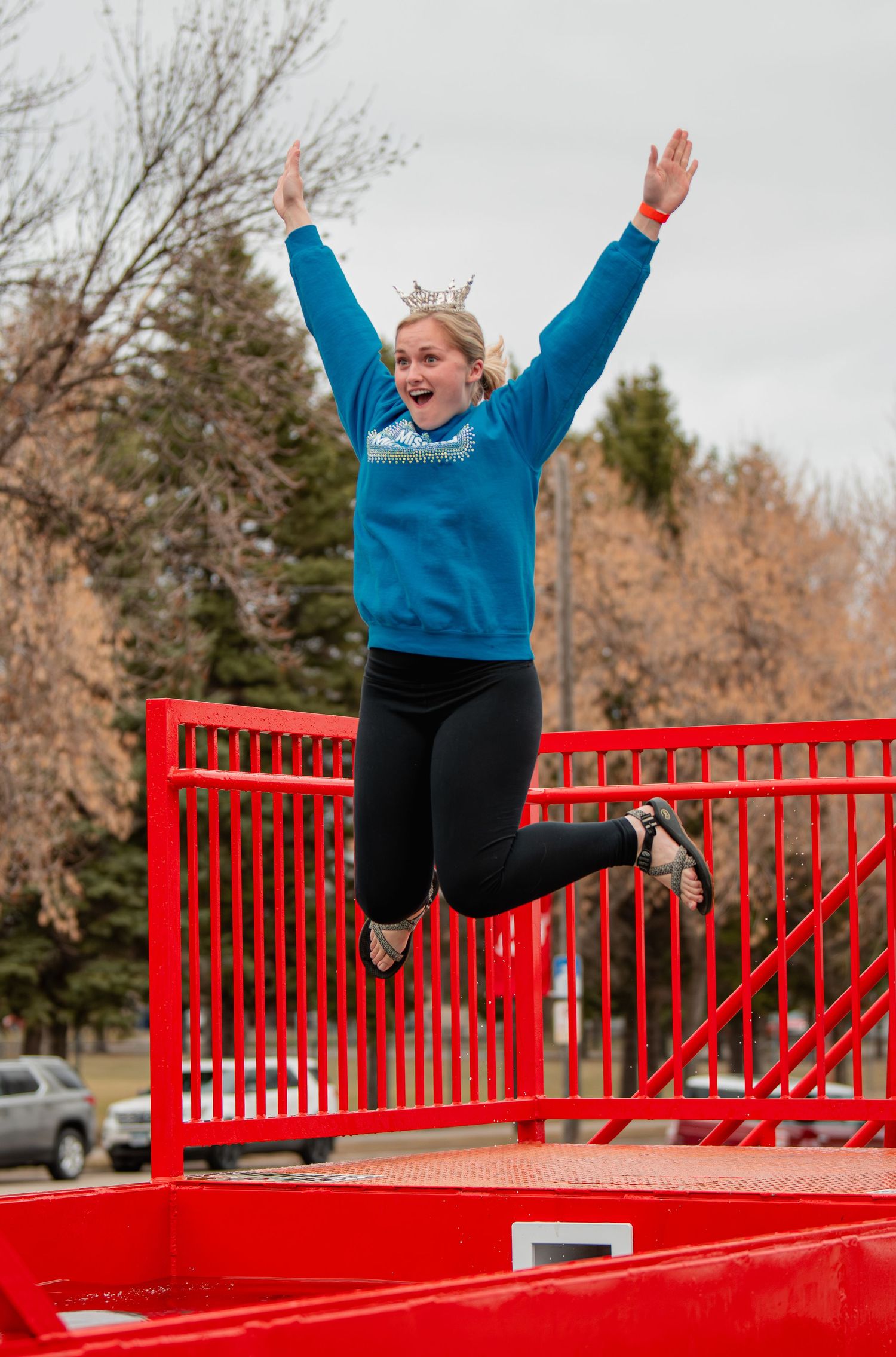 Girl wearing a crown jumps joyfully on a red trampoline platform outdoors, arms raised high, wearing a blue sweater and black pants.
