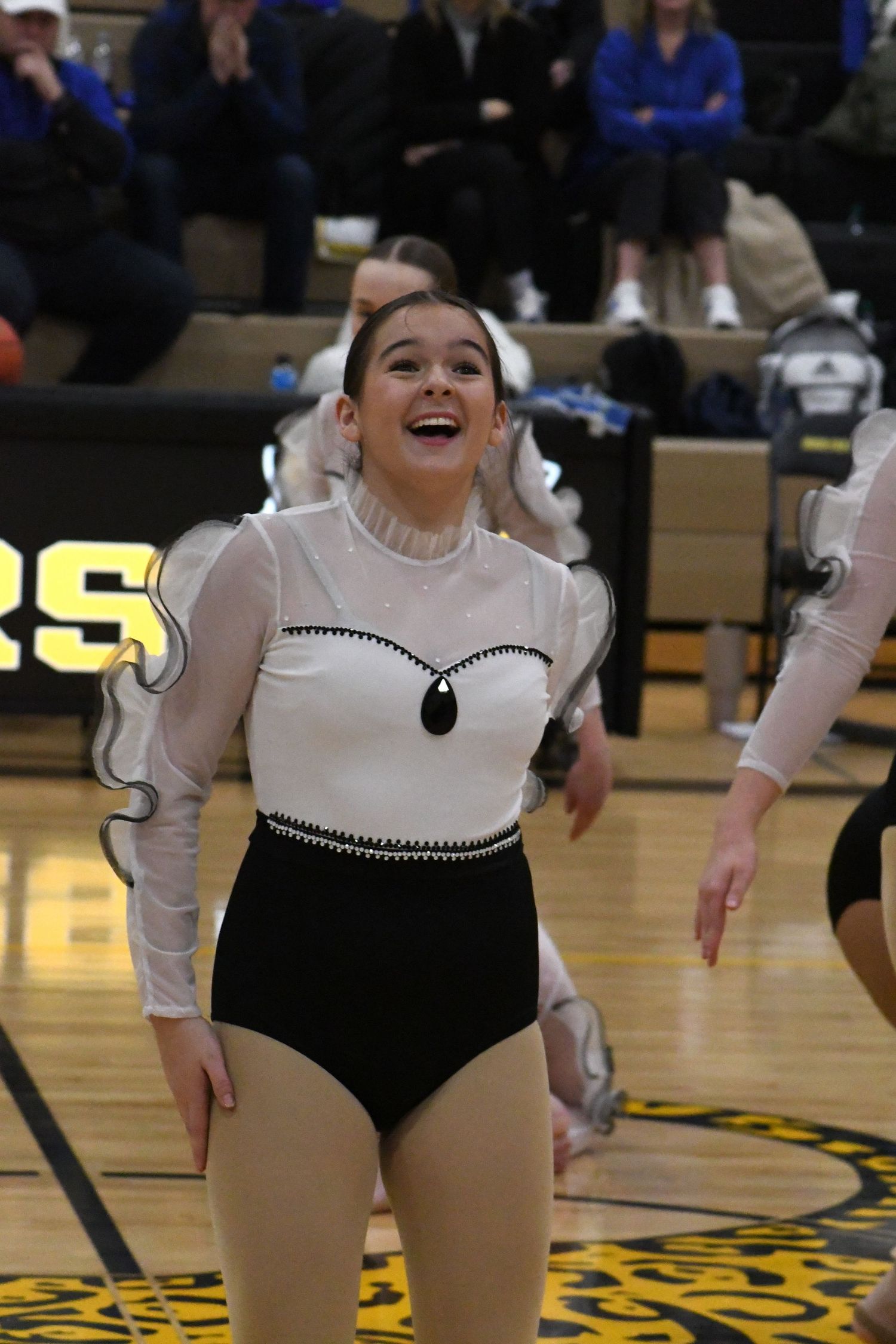 Performer in black and white leotard smiles during dance routine in school gymnasium.
