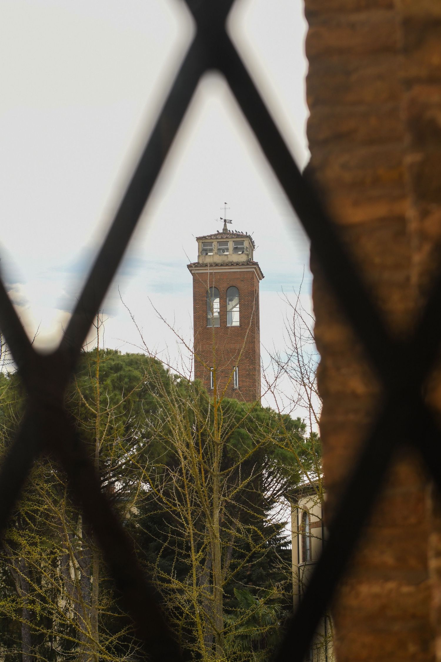 Historic brick tower with bell structure viewed through diamond-patterned iron fence.