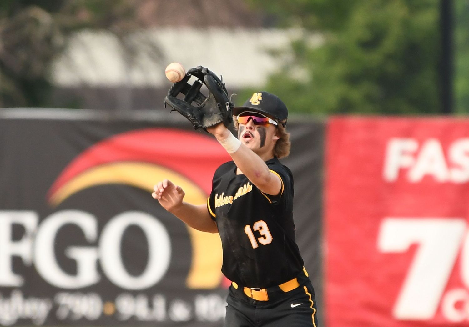 Baseball player in black and gold uniform reaches up to catch ball during game.