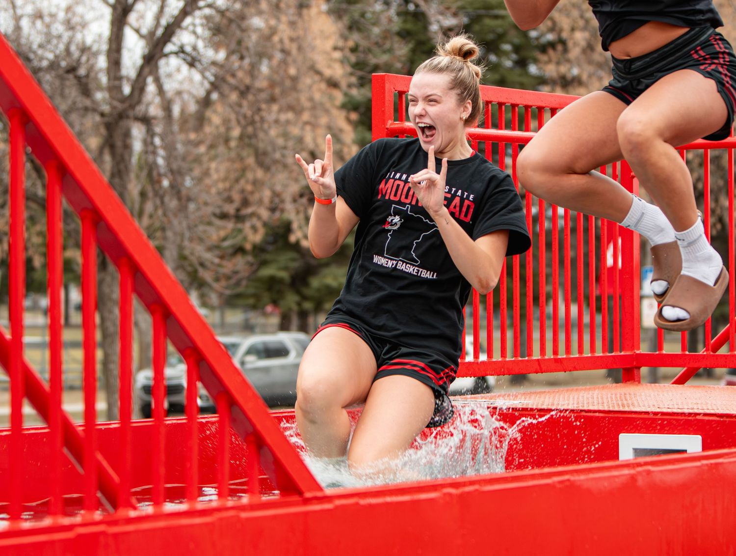 A young woman in a black shirt reacts with excitement as she gets splashed in a red dunk tank outdoors.