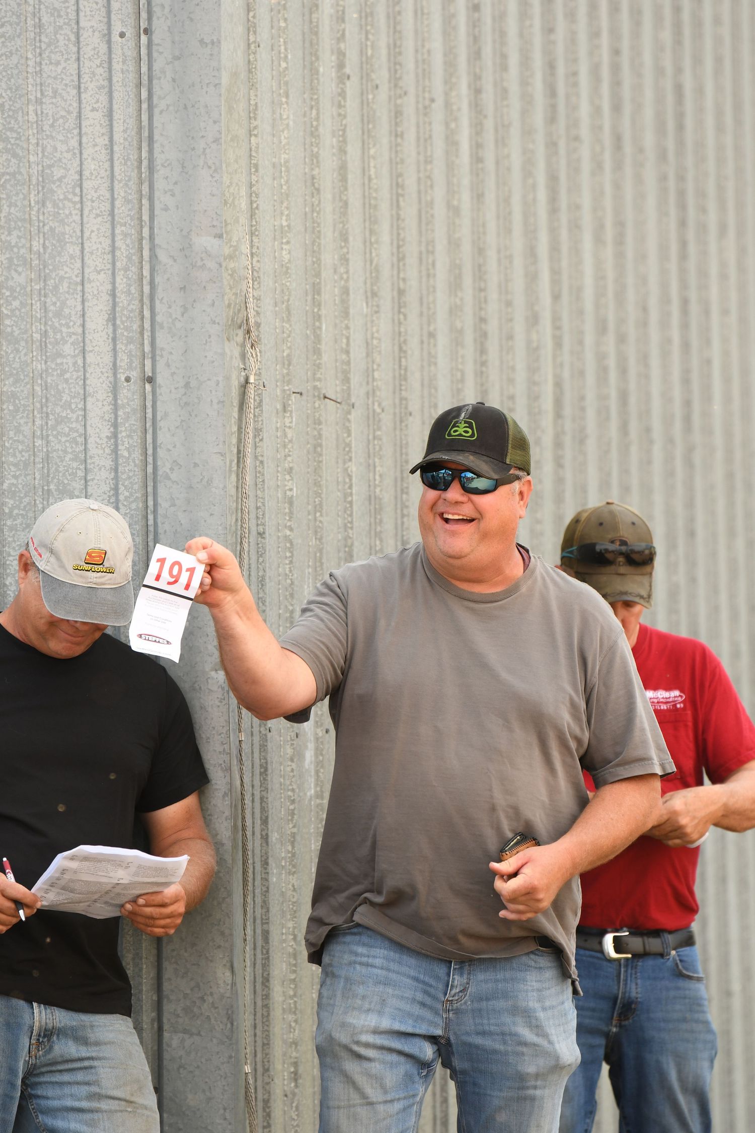 Three men stand against a metal wall checking documents, with one person in a gray shirt holding up paperwork.