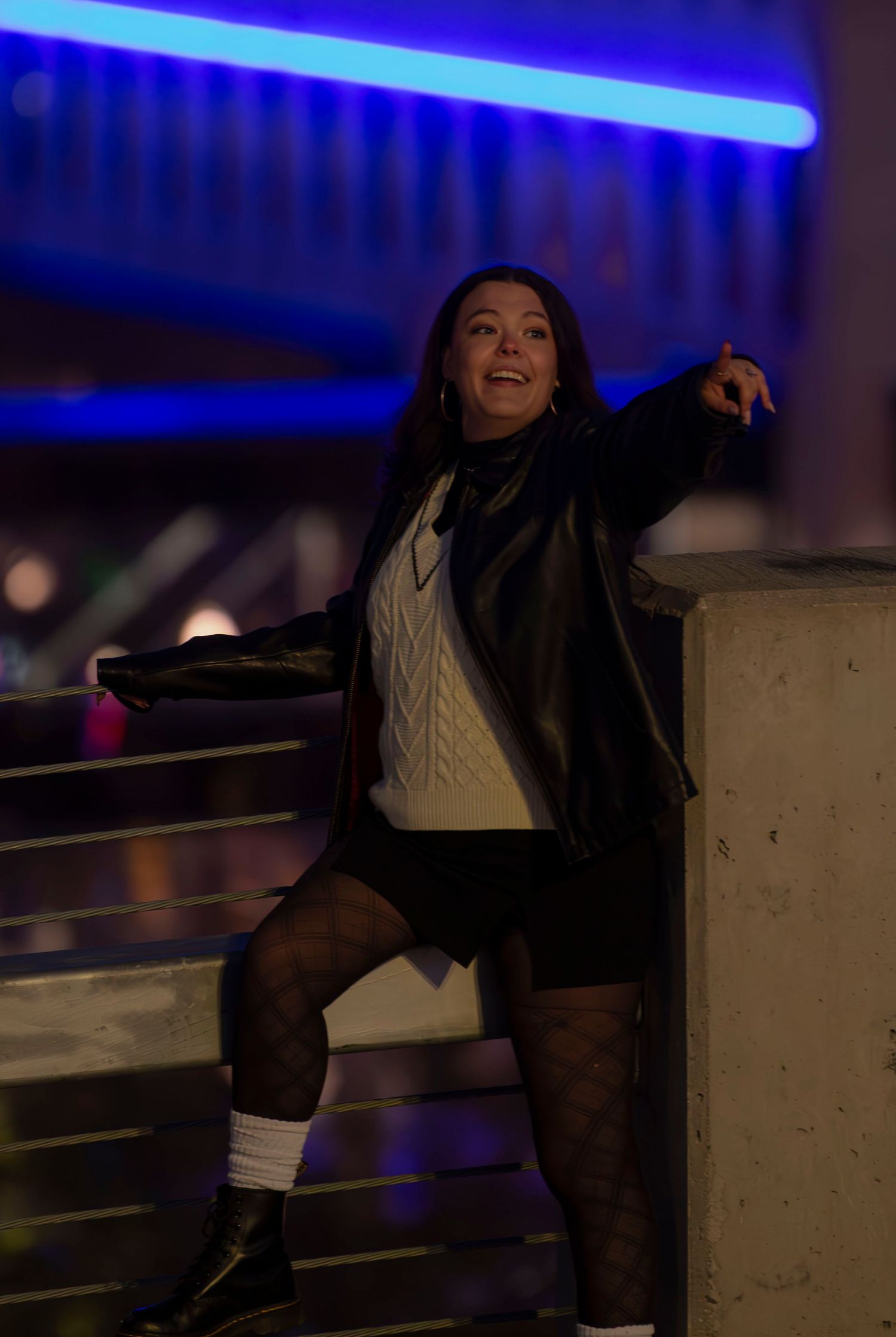 A dynamic nighttime pose under blue neon lighting with a leather jacket and patterned tights.