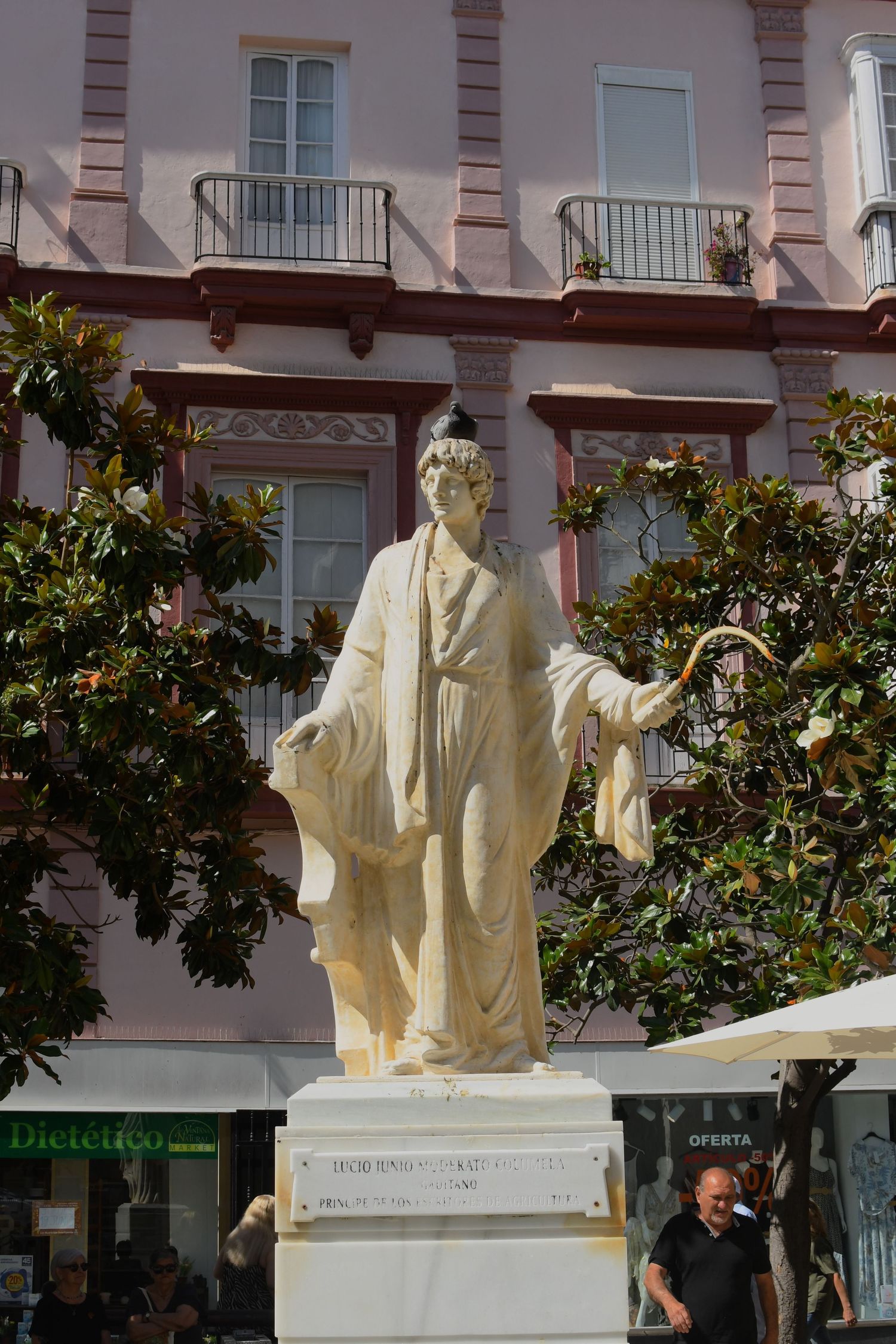 White marble statue in a classical pose stands in front of pink buildings with balconies.