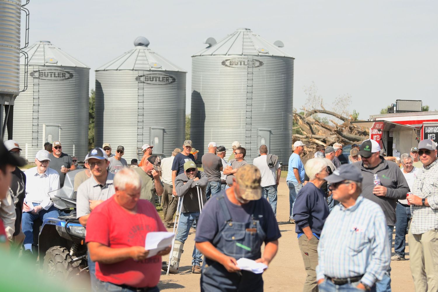 Large group of farmers and agriculture professionals gather around silver grain storage silos during a field day event.