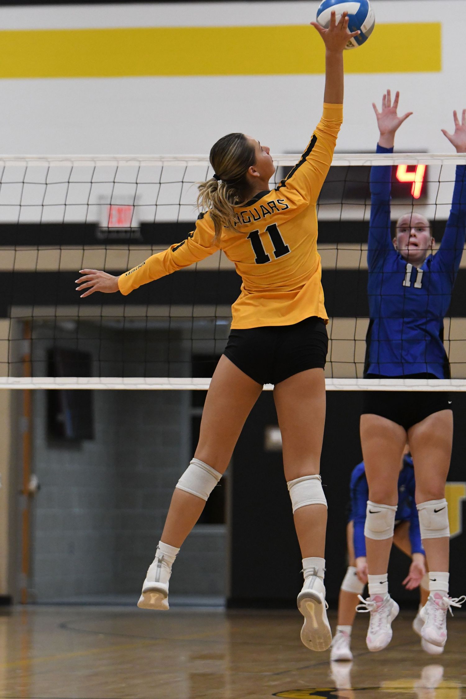 Player in yellow jersey stretches to spike volleyball over the net during match.