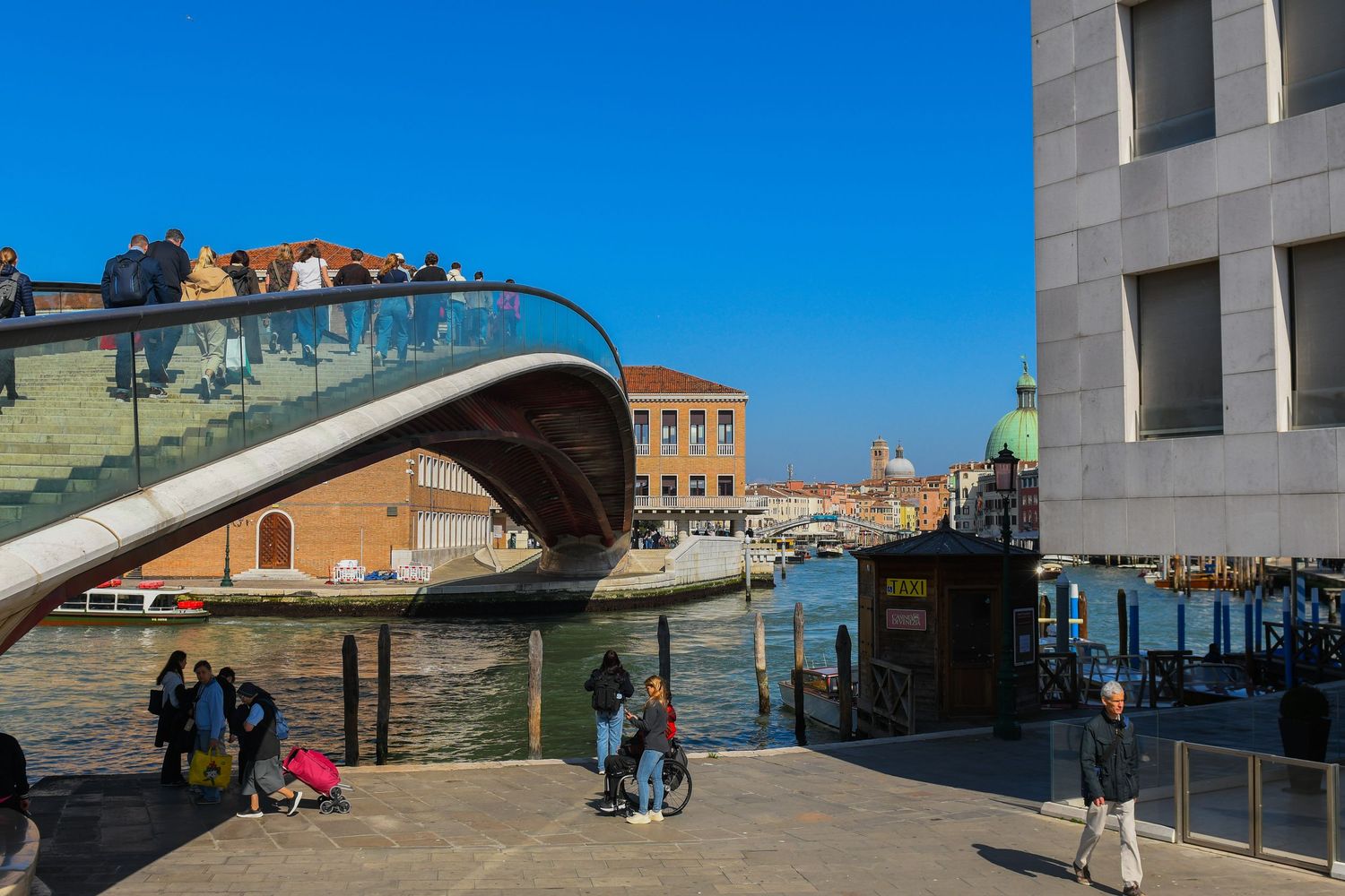 Modern glass bridge spans across a Venice canal with historic buildings in the background.
