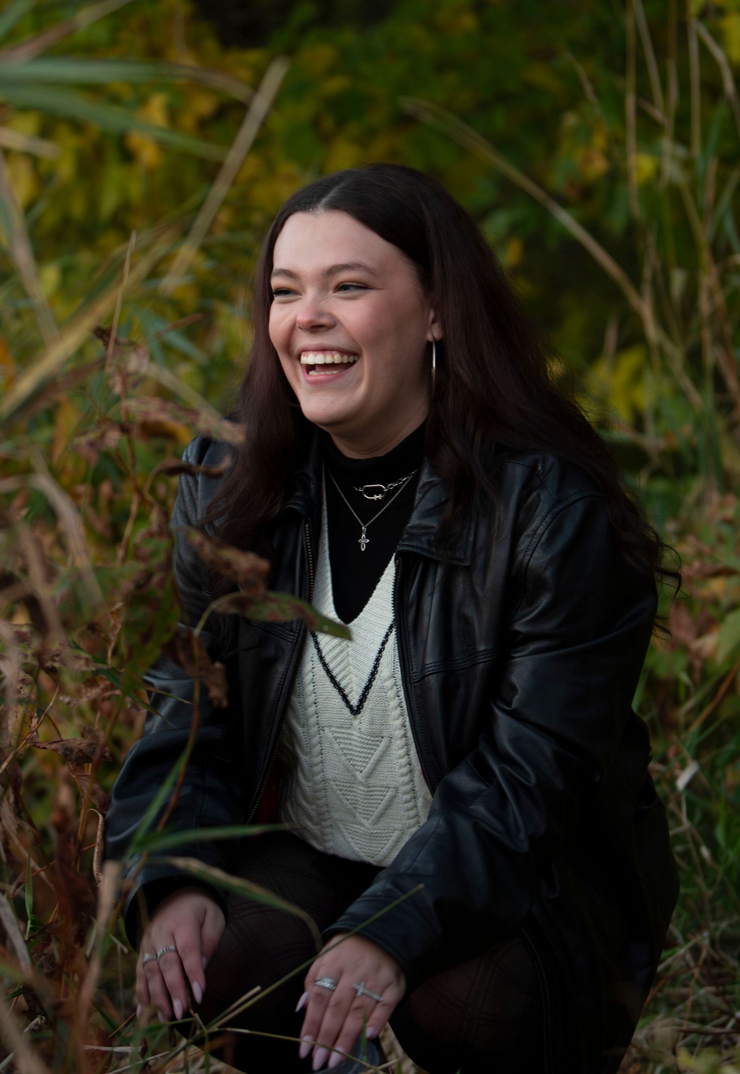 A joyful outdoor portrait featuring a white cable knit sweater and black leather jacket against natural foliage.