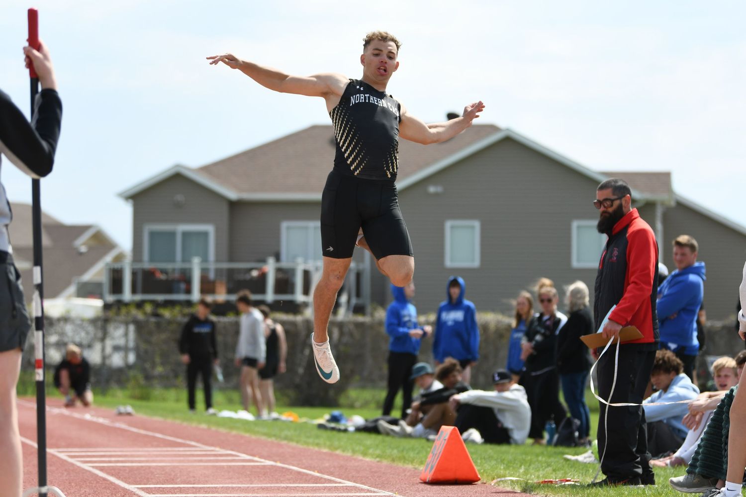 Athlete launches into air at track meet with houses visible in background.
