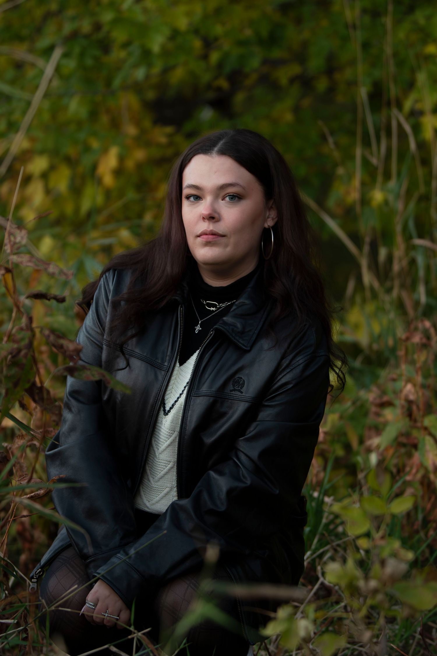 A person in a black leather jacket sits outdoors among autumn foliage in a moody natural setting.