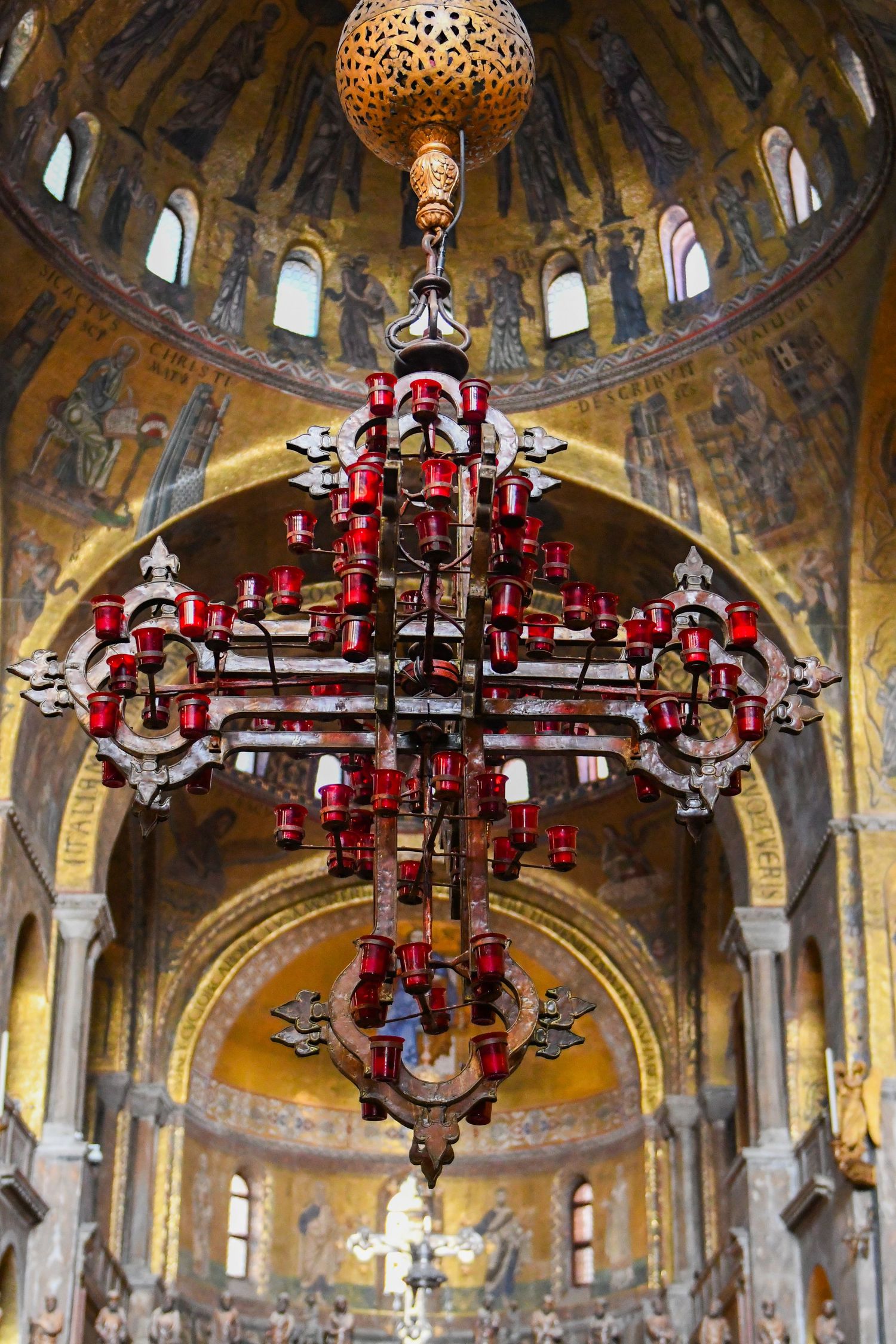 Ornate red and gold chandeliers hanging in a church with Byzantine-style mosaic ceilings.