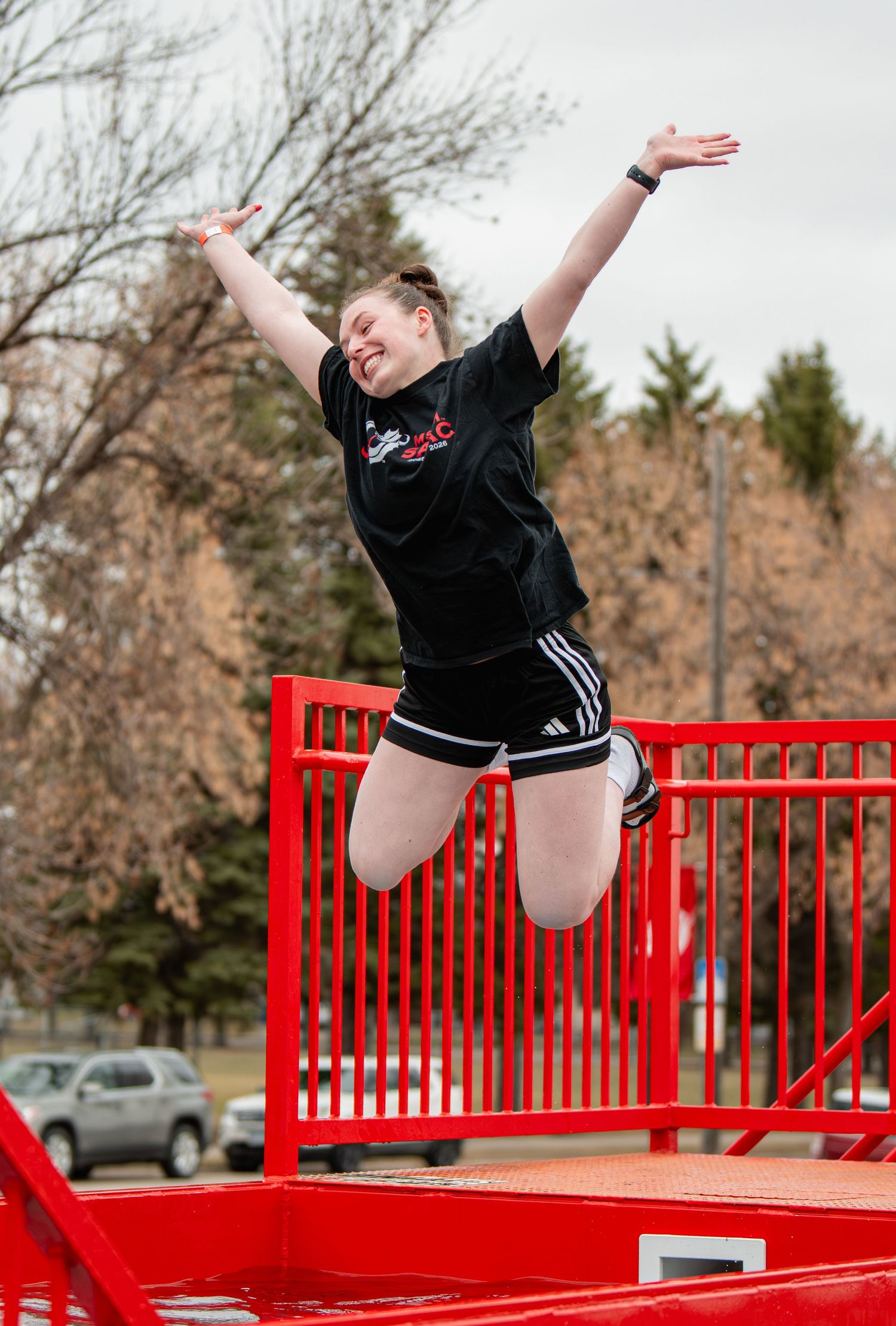 Young woman joyfully jumping in the air outdoors near a red railing, wearing Adidas shorts and a black t-shirt.