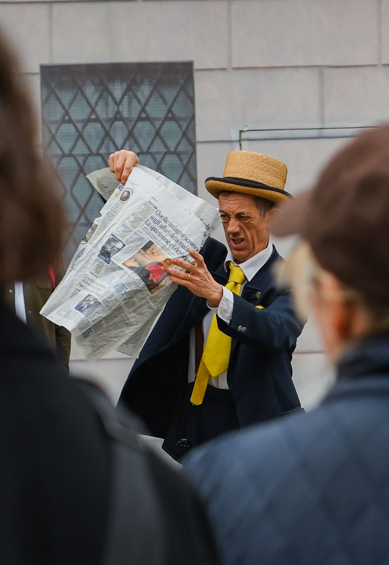 Person in hat and yellow scarf gestures while holding newspapers on city street.