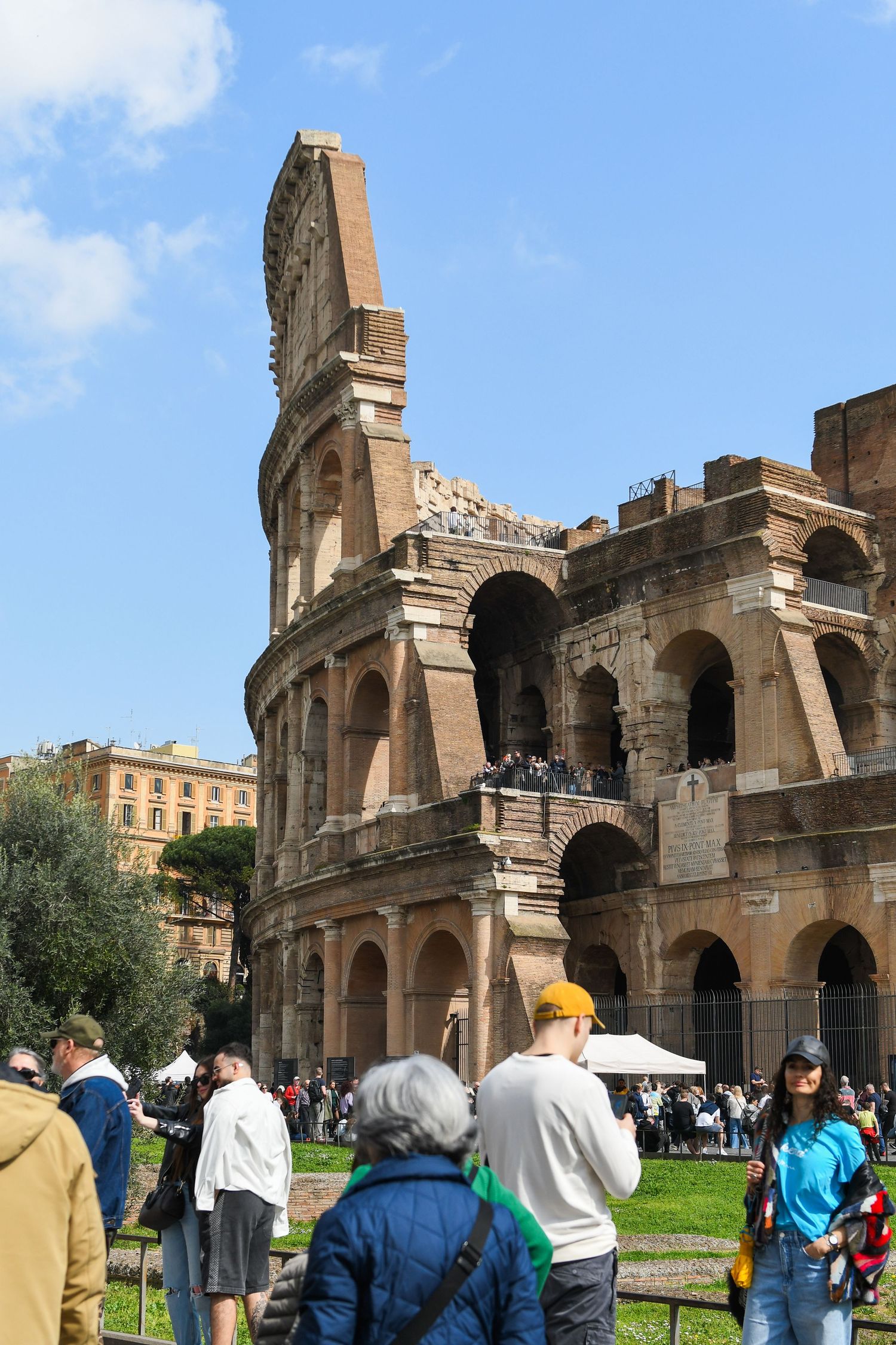 Exterior view of the Roman Colosseum with tourists gathering on the grass in front of the ancient amphitheater.