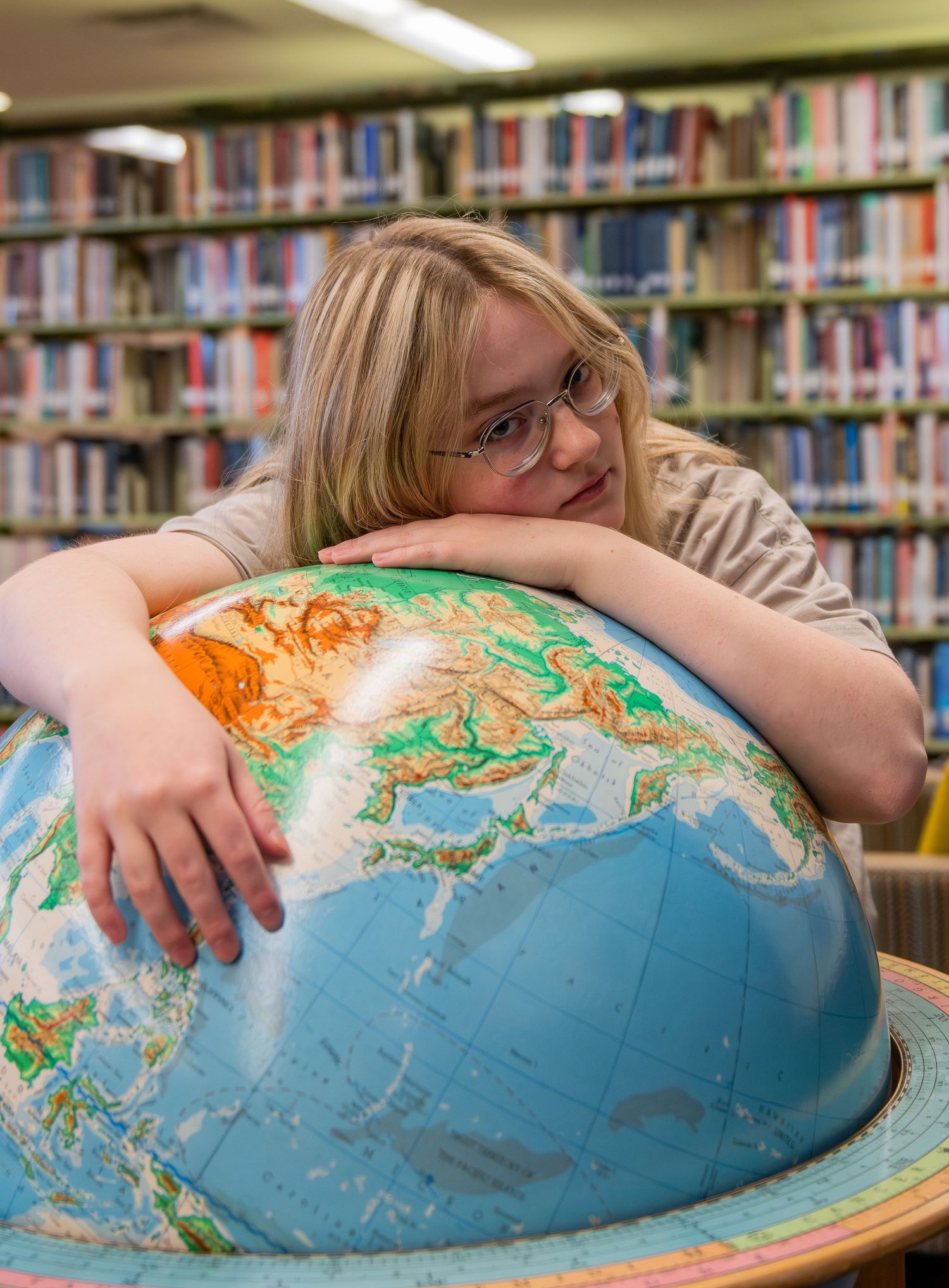 A person rests their head on a large globe while studying in a library surrounded by bookshelves.