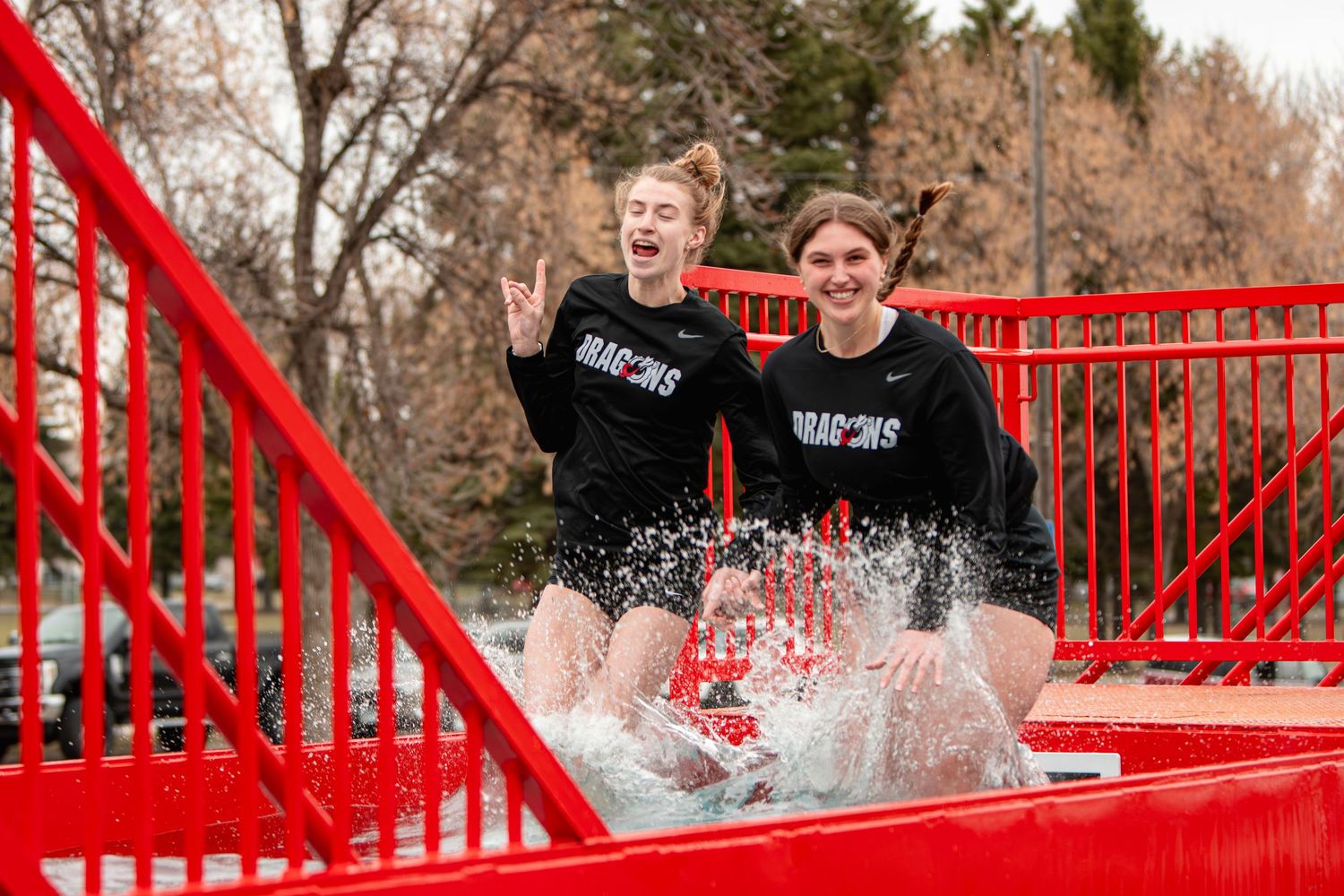 Two students in Dragons shirts laugh while sliding into icy water during a winter plunge event on a red slide.