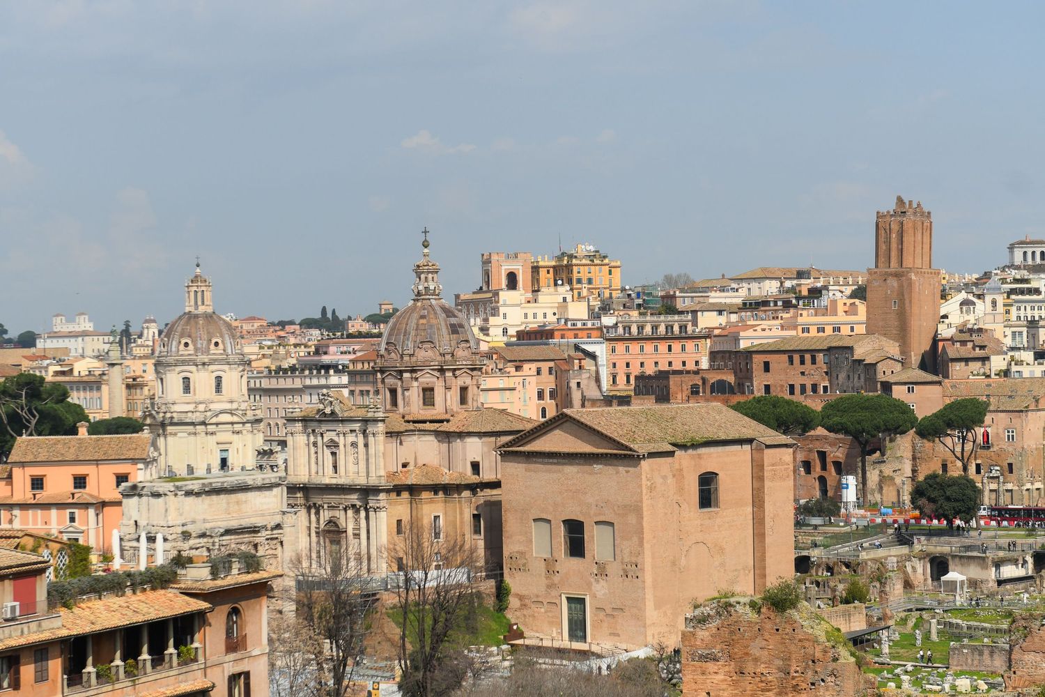 Panoramic view of Rome's historic skyline showing church domes, towers and terracotta rooftops against a blue sky.