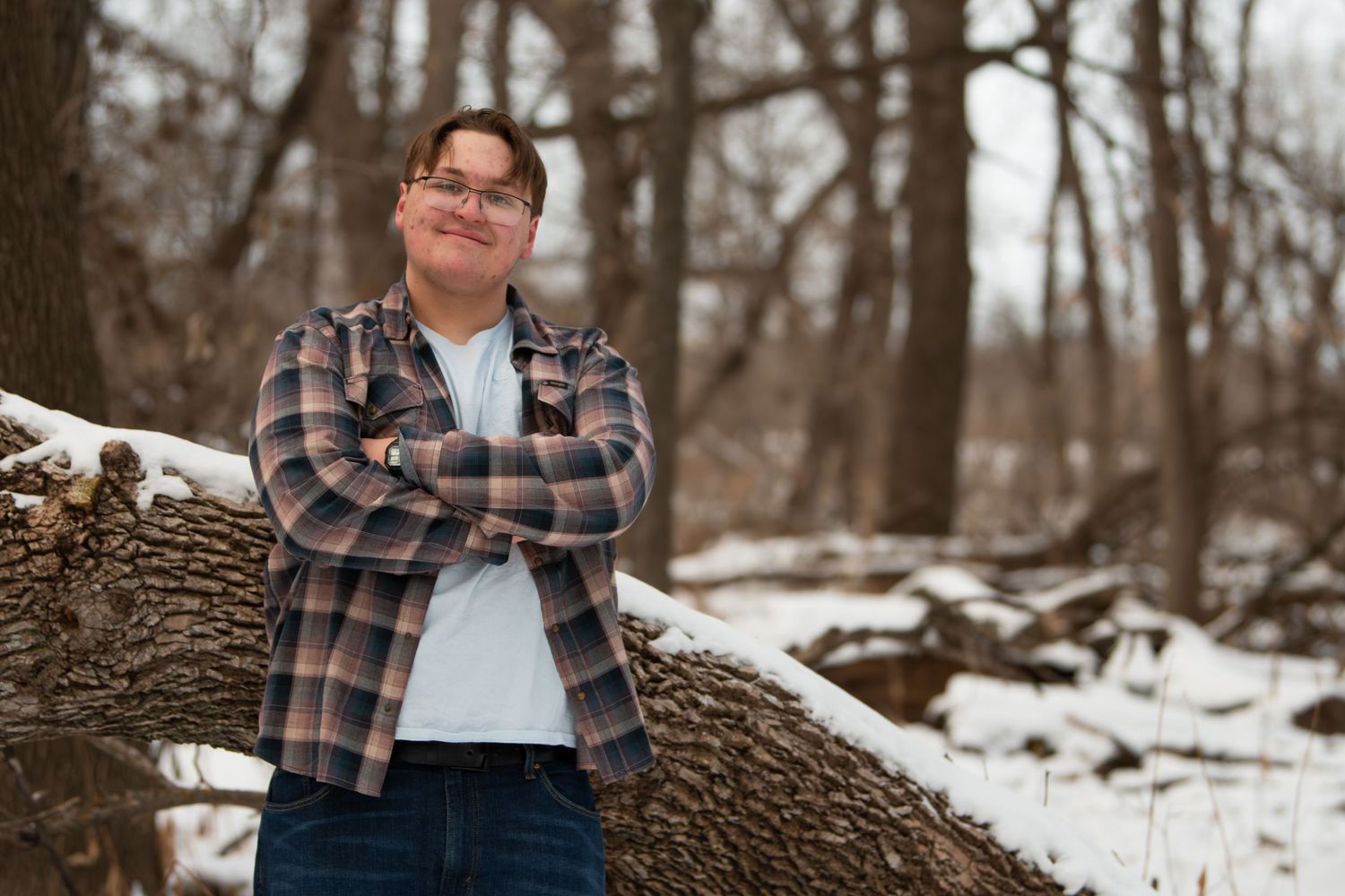A person wearing a plaid flannel jacket sits on a snowy log in a winter forest setting.