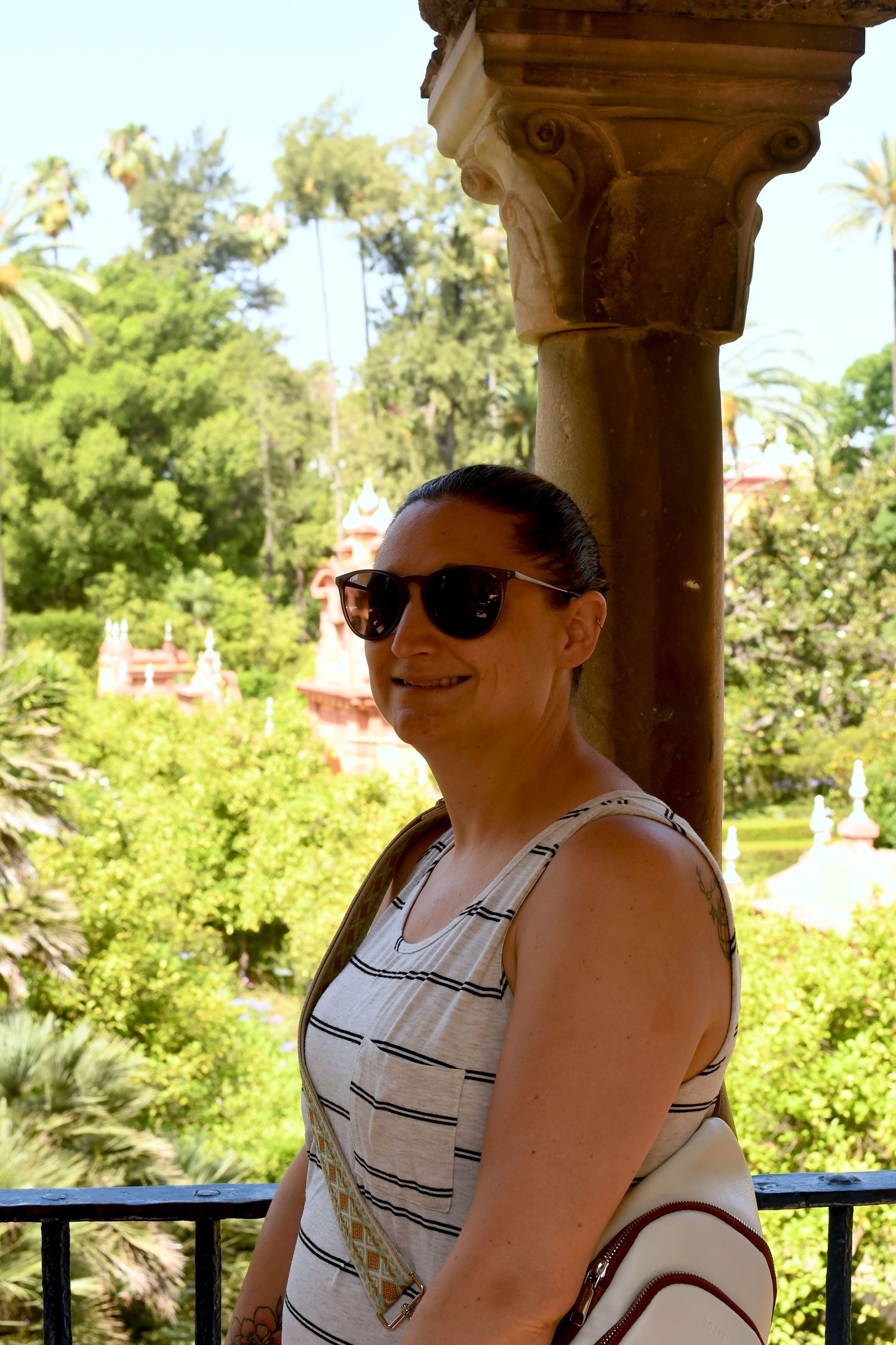 Tourist enjoying scenic view from ornate stone column balcony with tropical gardens in background.